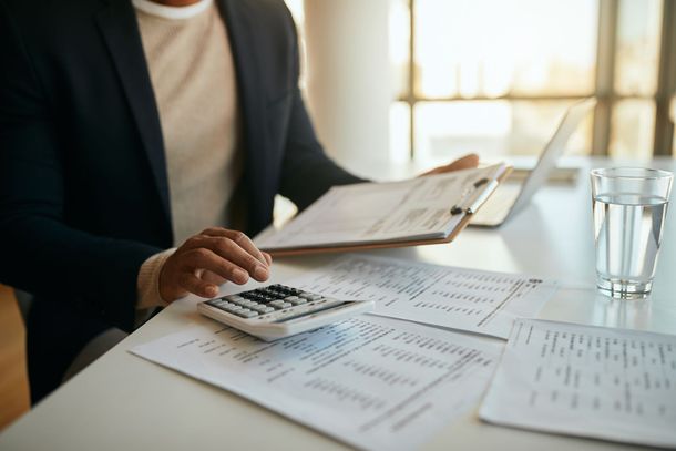 Person in blazer using calculator, reviewing paperwork, and near a laptop and a glass of water.