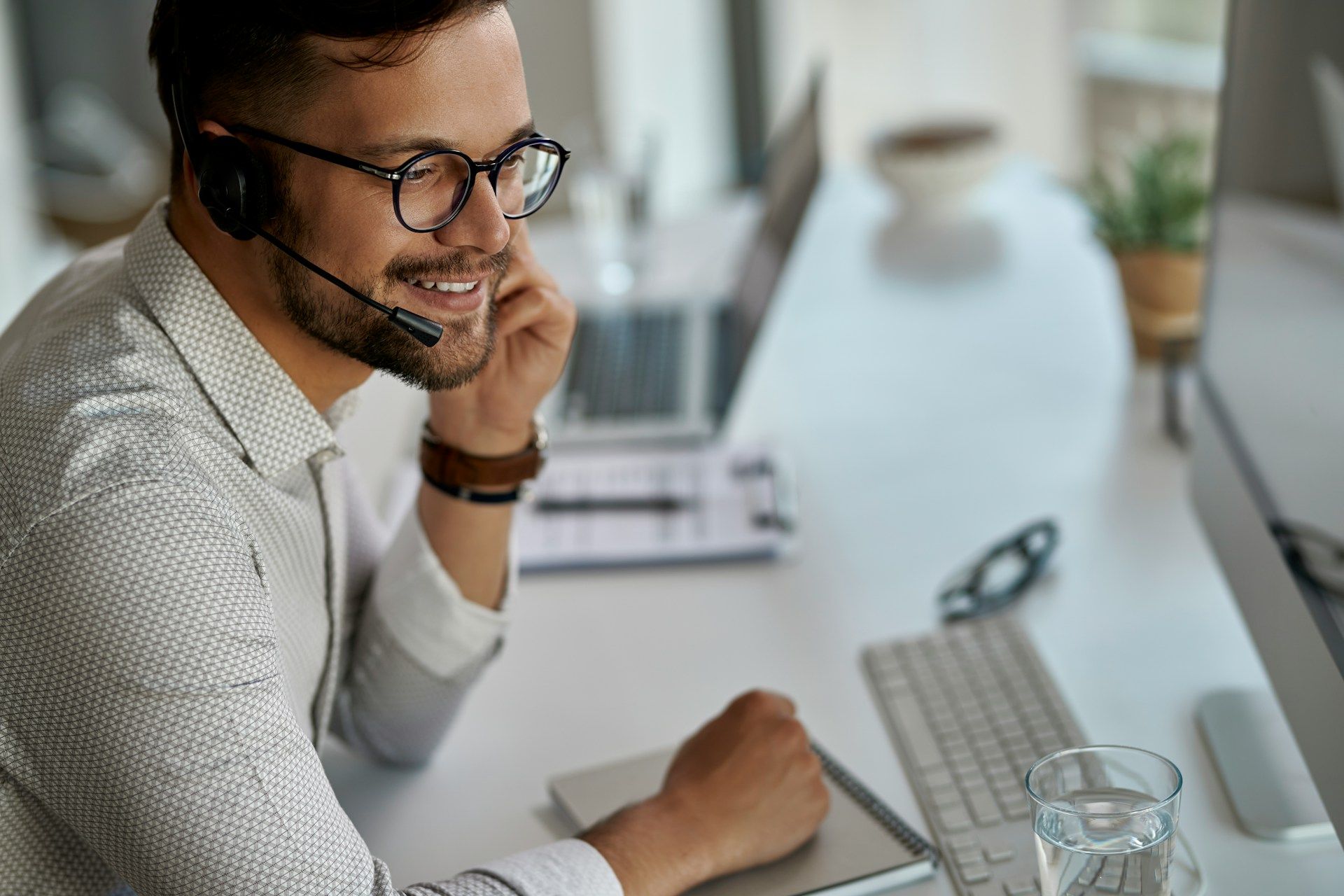 Man with glasses and headset smiling at computer, in an office setting.