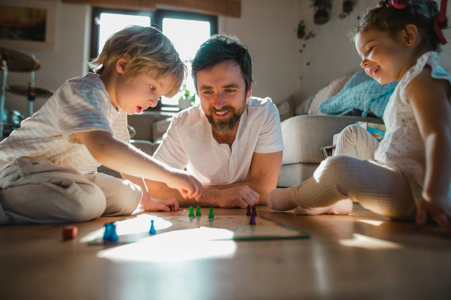 A man plays a board game with two young children on a wooden floor in a sunny room.