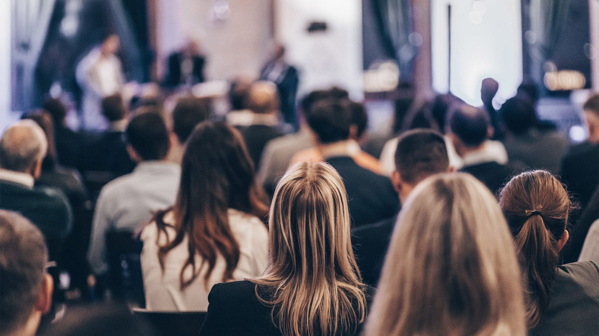 Audience seated at a conference, facing a stage with presenters; dim lighting.