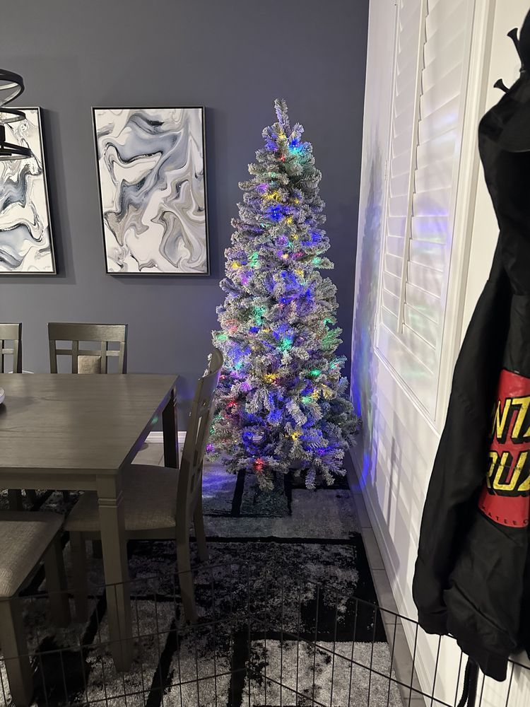 A tall, flocked Christmas tree with colorful lights stands in a dining area next to a window.