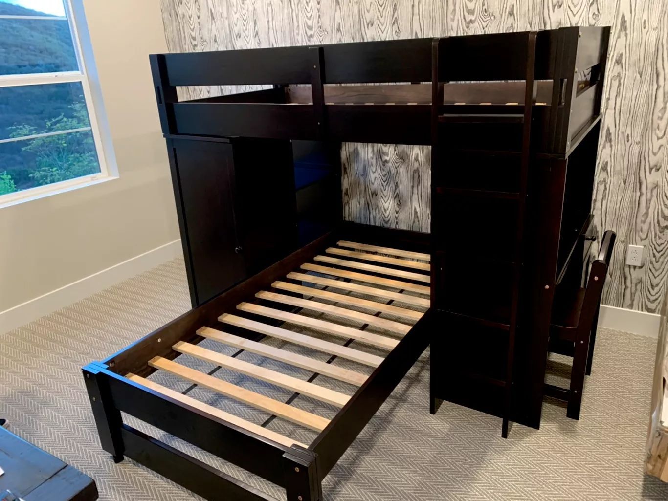 Dark bunk bed with a pull-out bed, desk, and storage against a patterned wall, by a window.