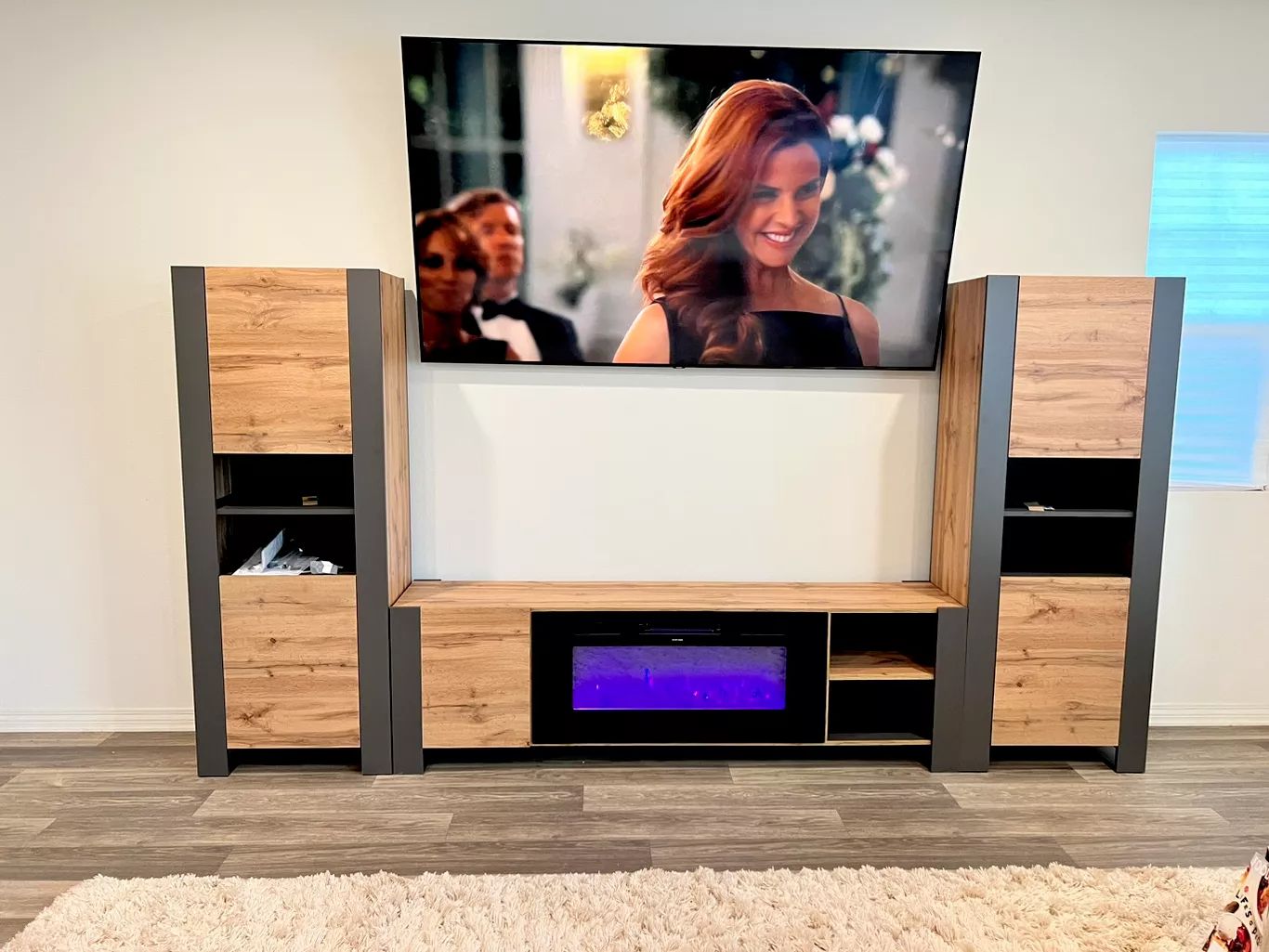 A wall-mounted TV above a gray and brown entertainment center. The TV displays a woman smiling.