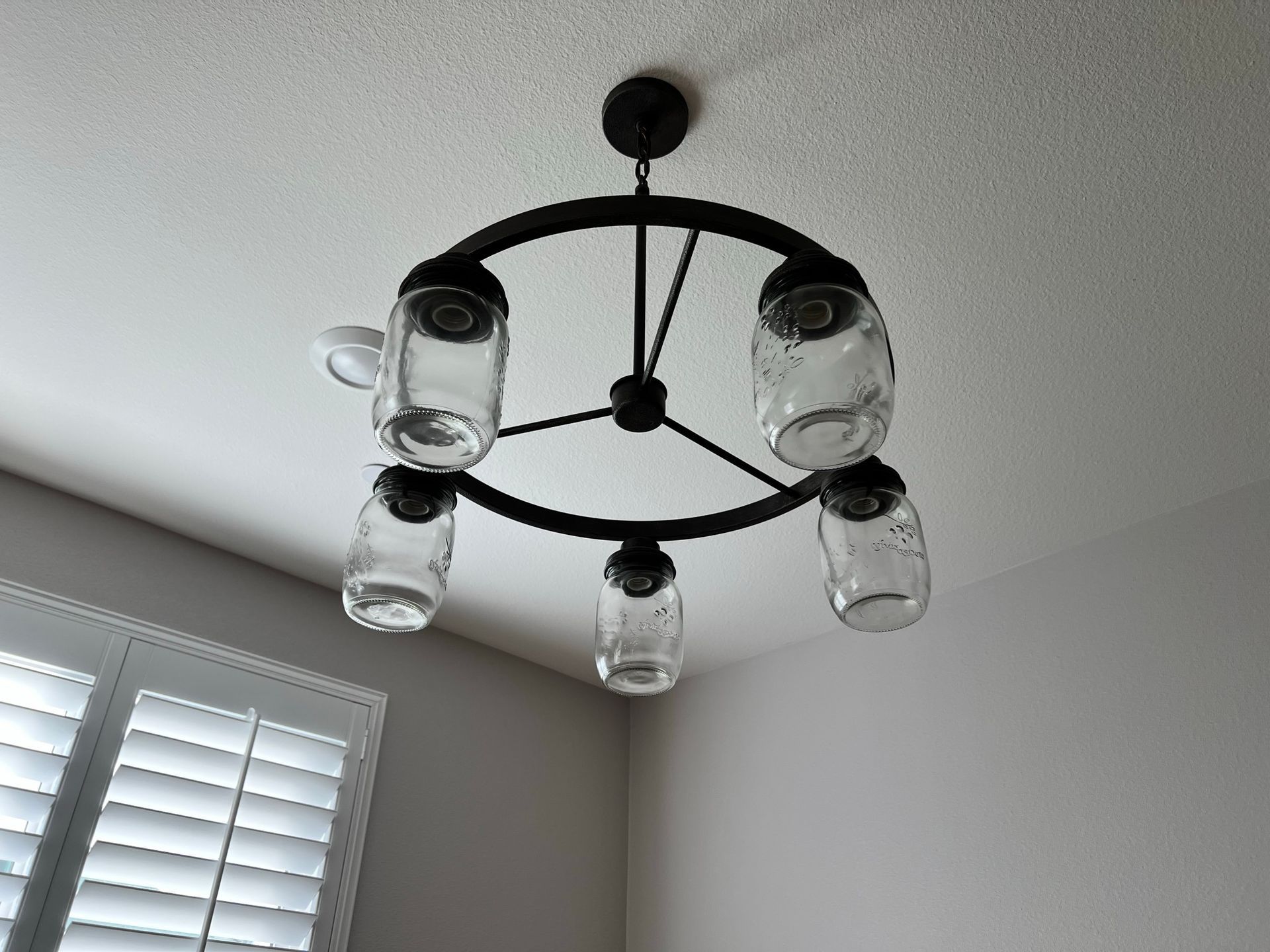 A chandelier with five glass jar lights hangs from a white ceiling, near a window.