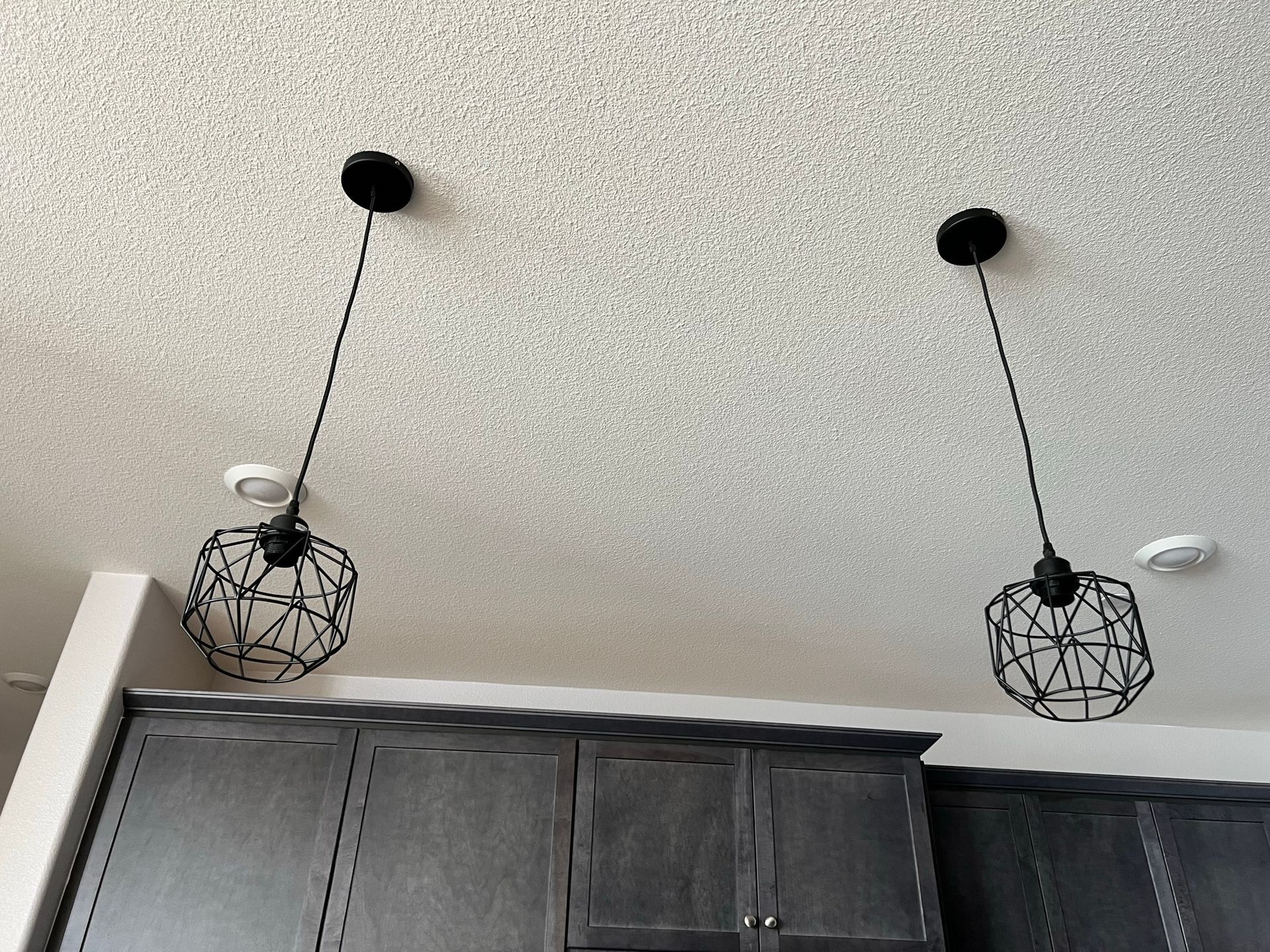 Two pendant lights with black wireframe shades hanging above dark gray kitchen cabinets.