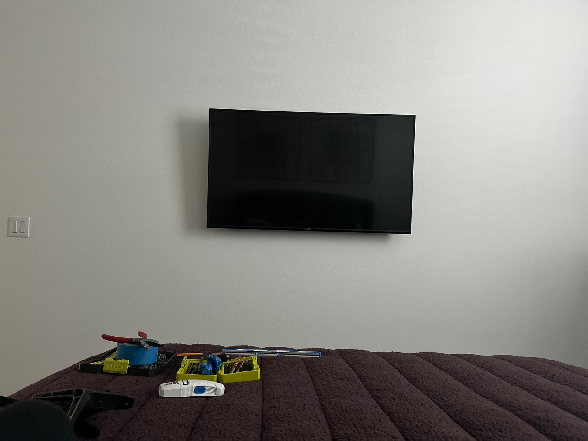 A wall-mounted TV above a bed with toys on it. The wall is white.