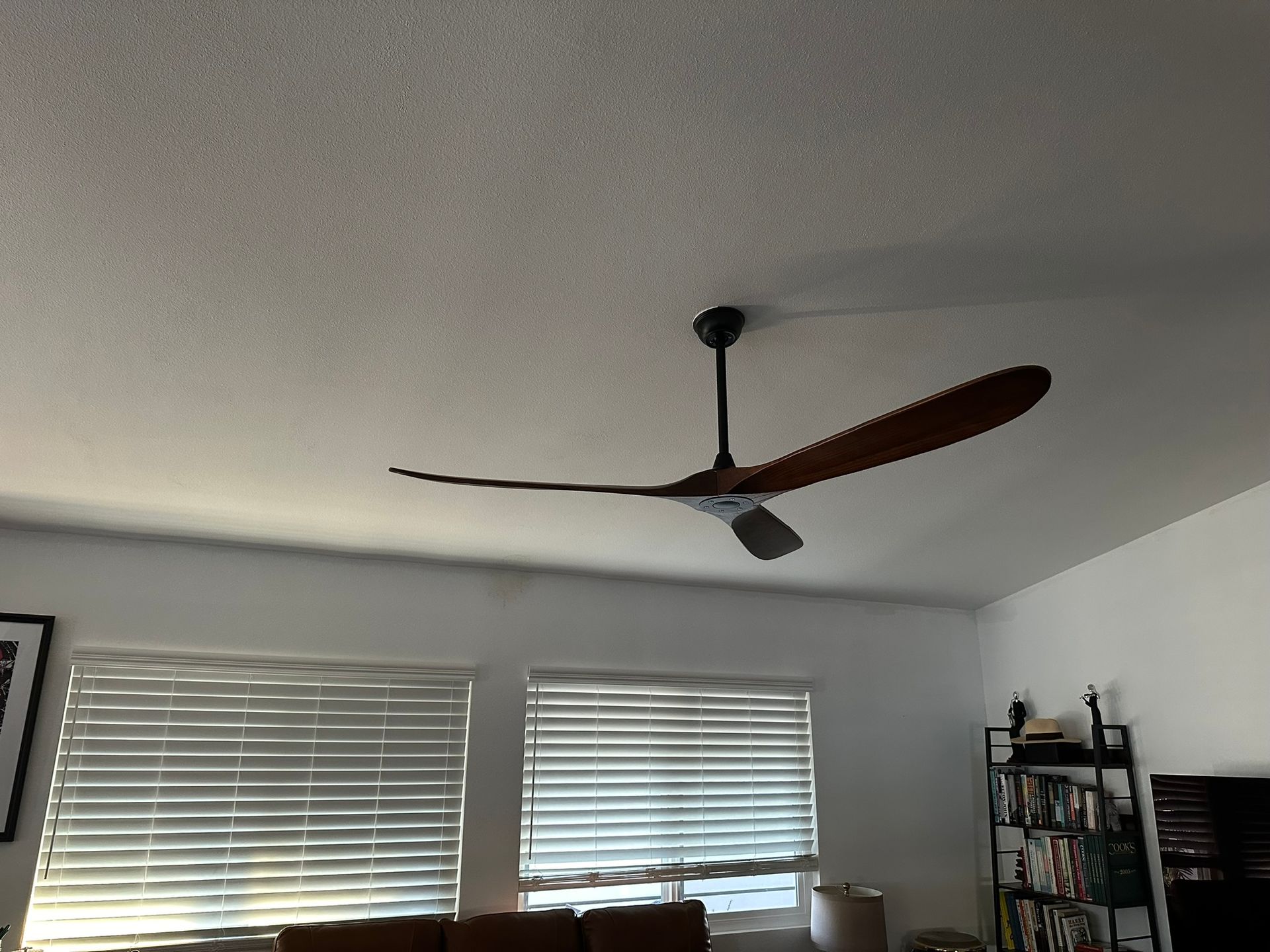 Ceiling fan with wooden blades in a room with blinds, shadows, and bookshelves.
