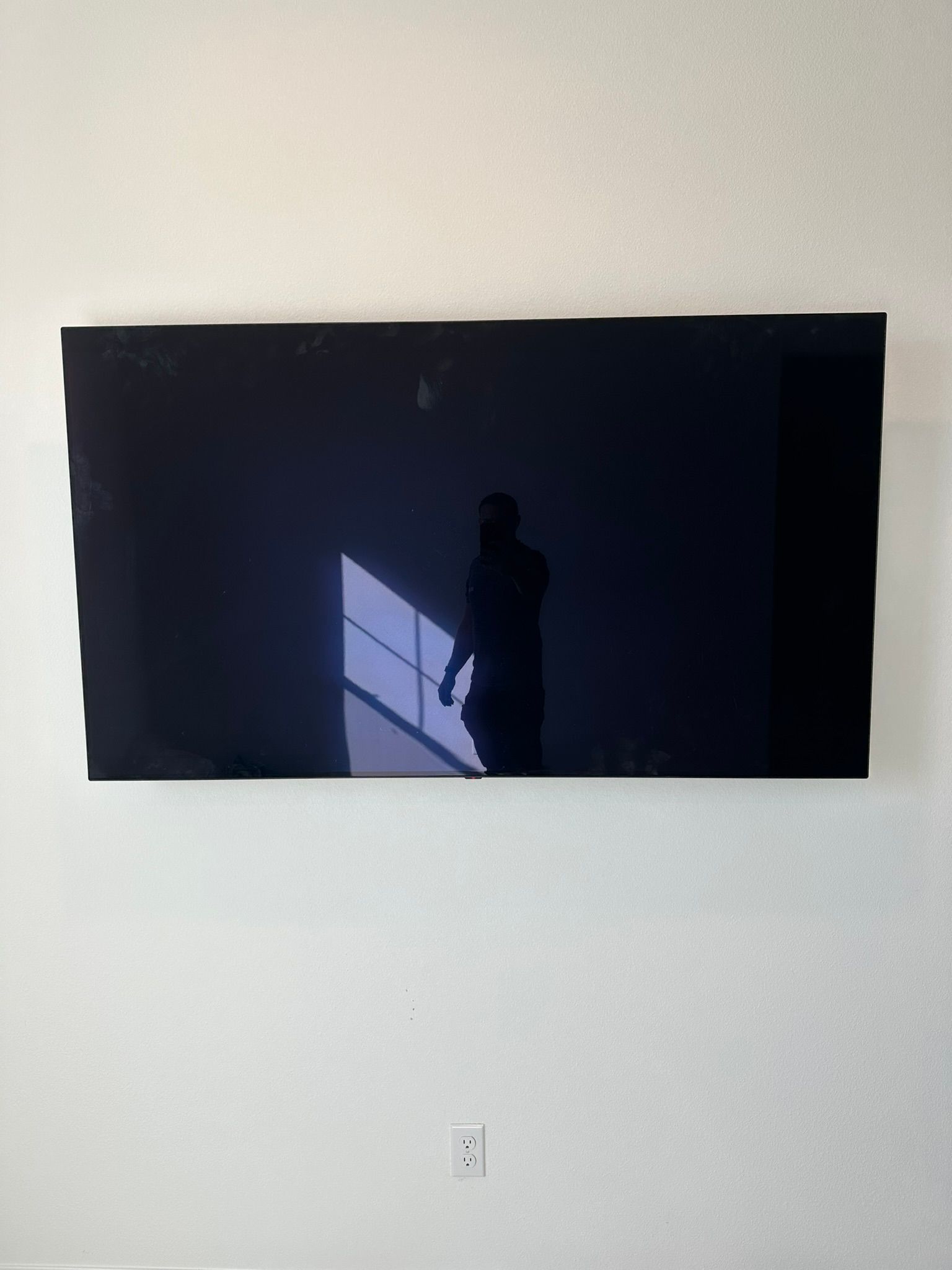 Large, black, rectangular television mounted on a white wall. Reflected in screen: person and window with sunlight.