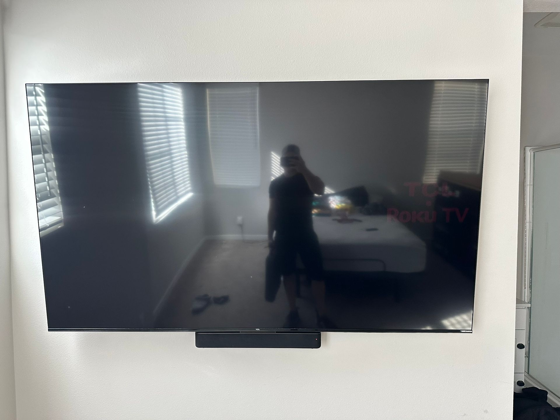 Large TV mounted on a white wall, reflecting a person holding a phone in a room with a bed and window.