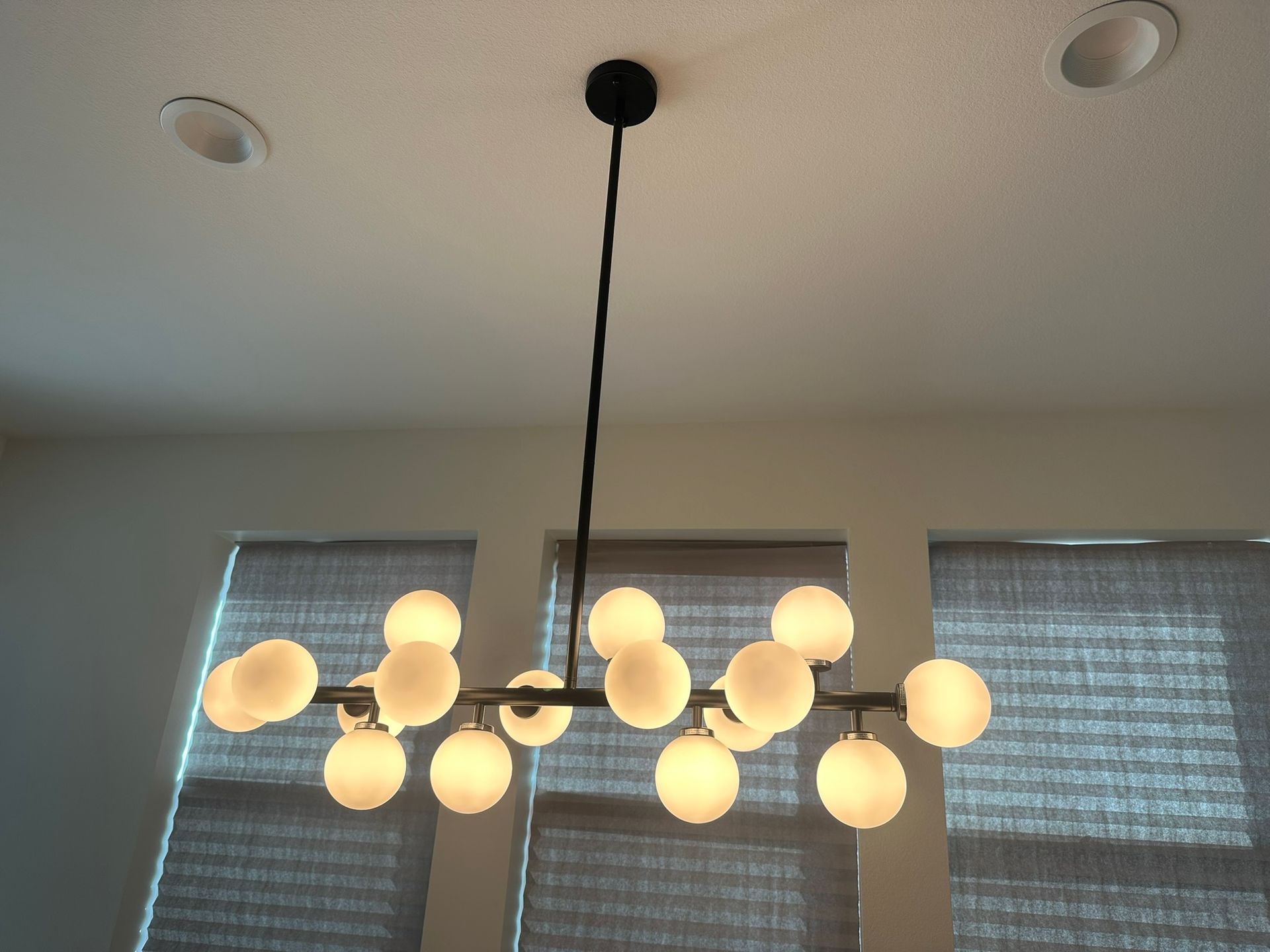 Modern chandelier with spherical lights hanging from a black rod, near windows with gray blinds.