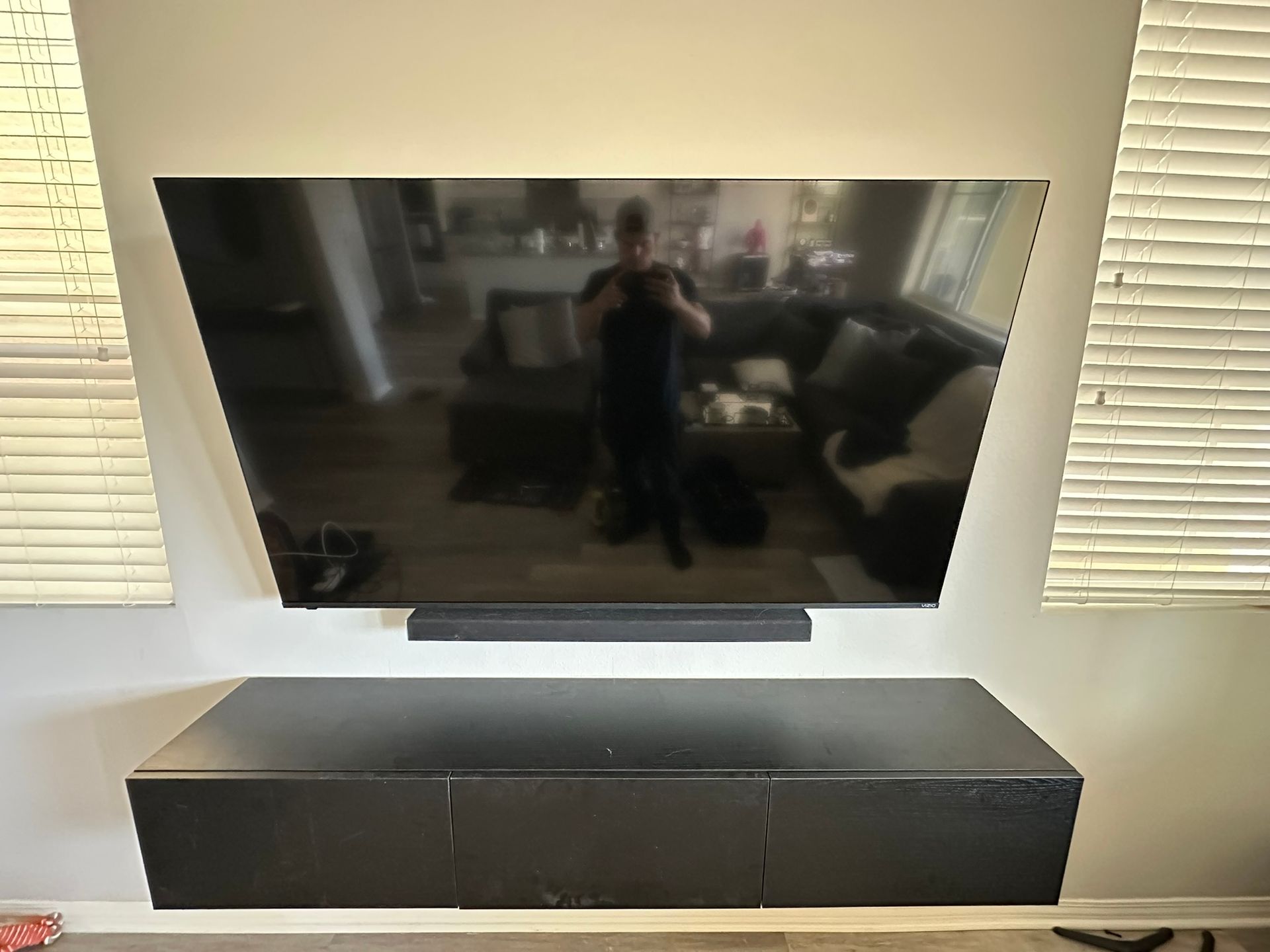 Wall-mounted TV with soundbar above a black cabinet, the reflection showing a person in the living room.