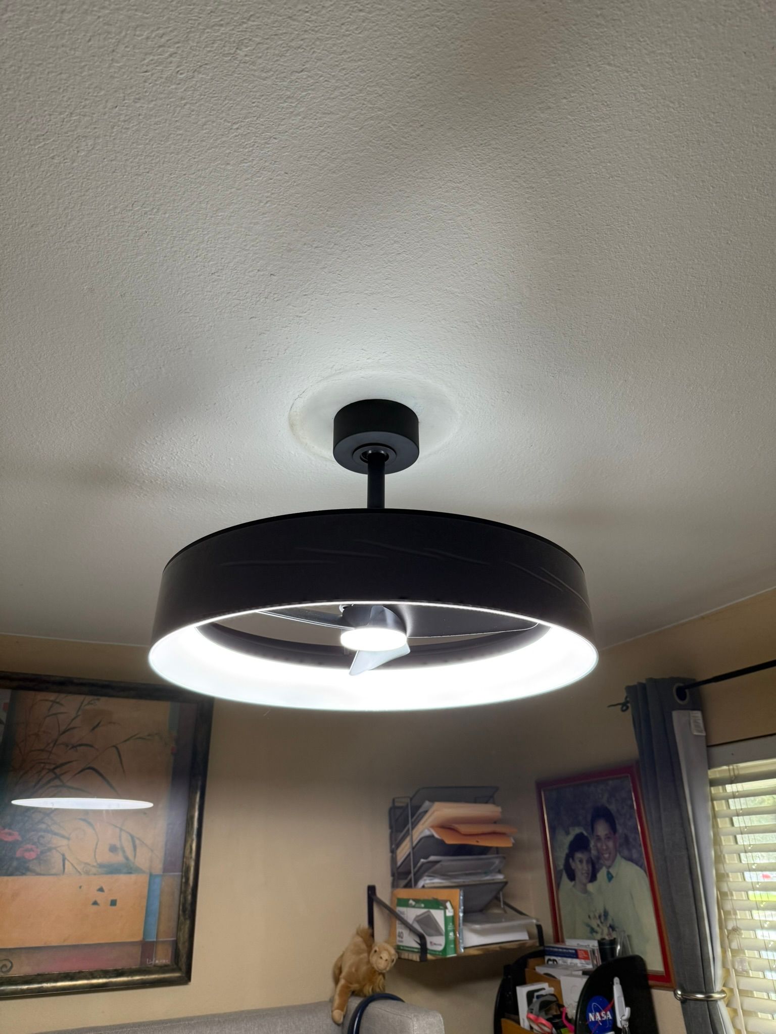 Black ceiling fan with a bright circular light, mounted on a textured ceiling in a room with artwork and curtains.