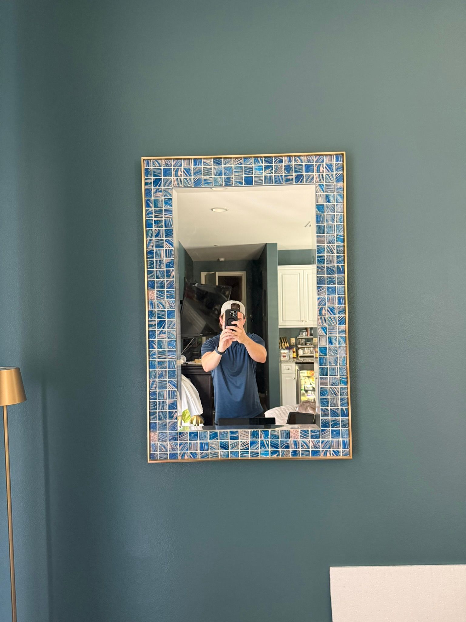 Mirror with blue mosaic frame reflecting a person taking a photo in a teal-walled room