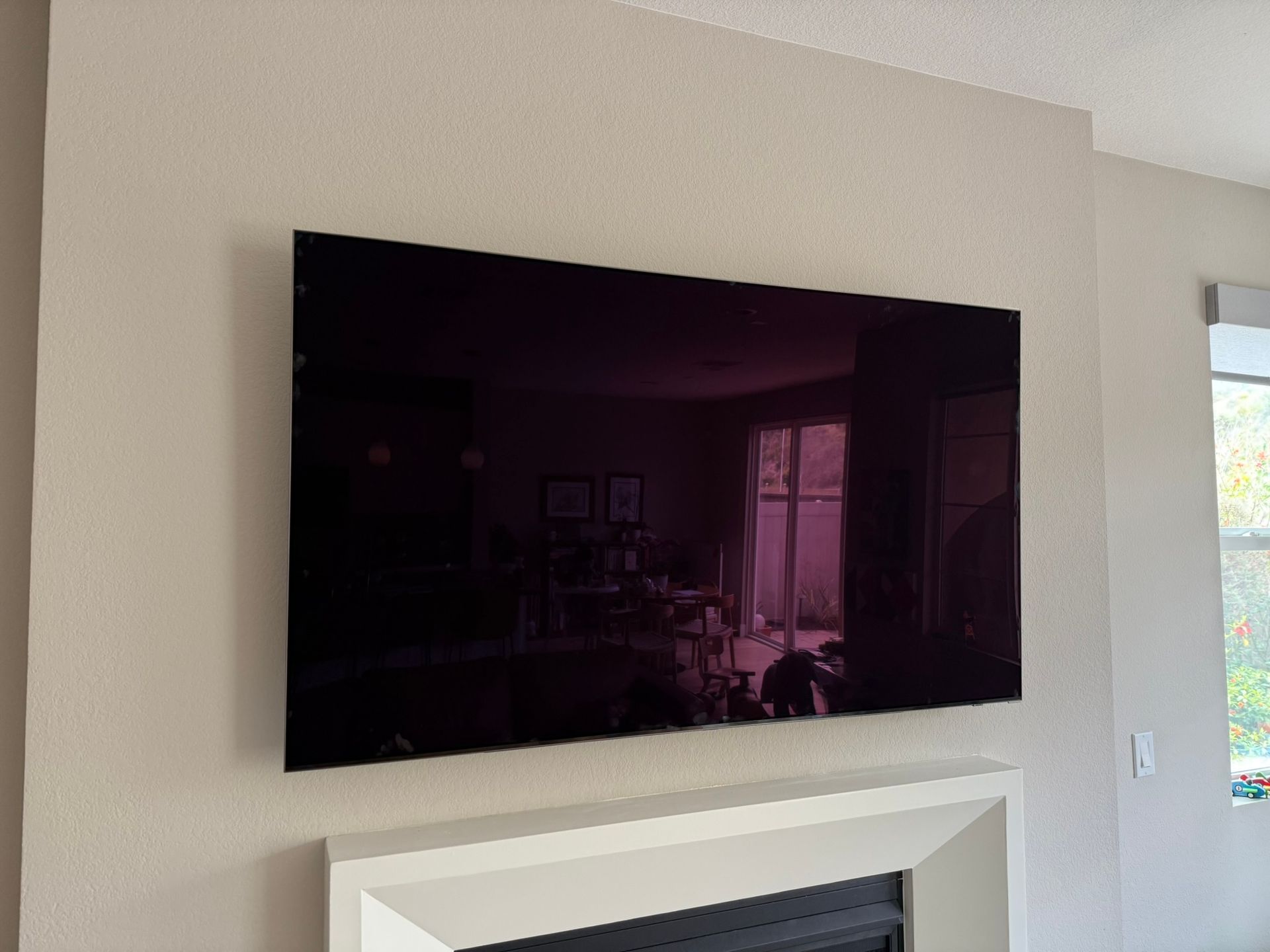 Large flat-screen TV mounted above a white fireplace. The screen reflects a room with windows.