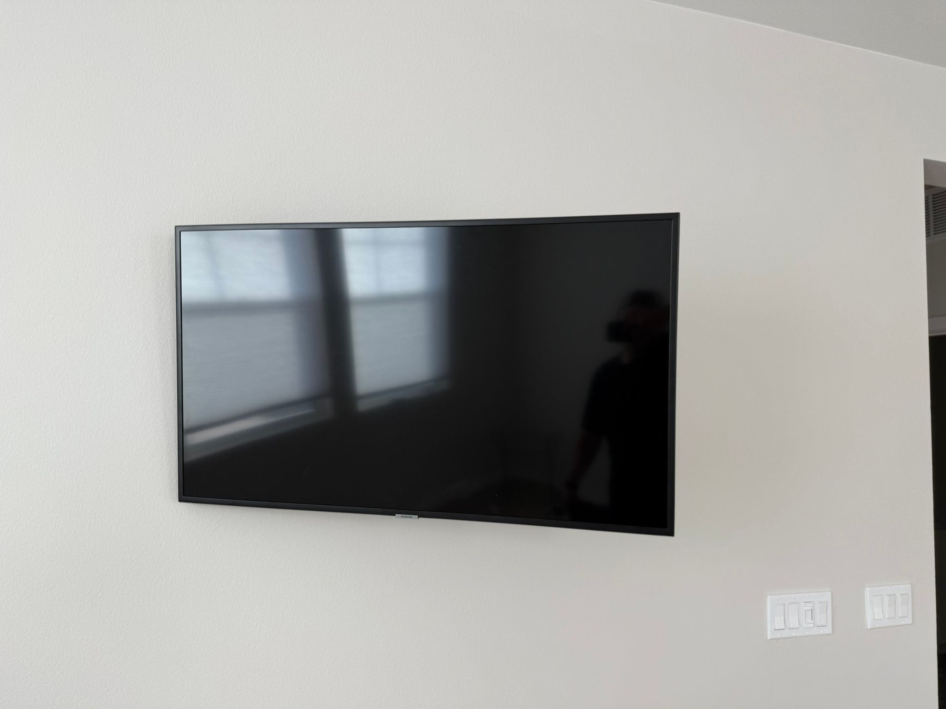 A large flat-screen TV mounted on a light beige wall. Reflected in the screen is a window and a person.