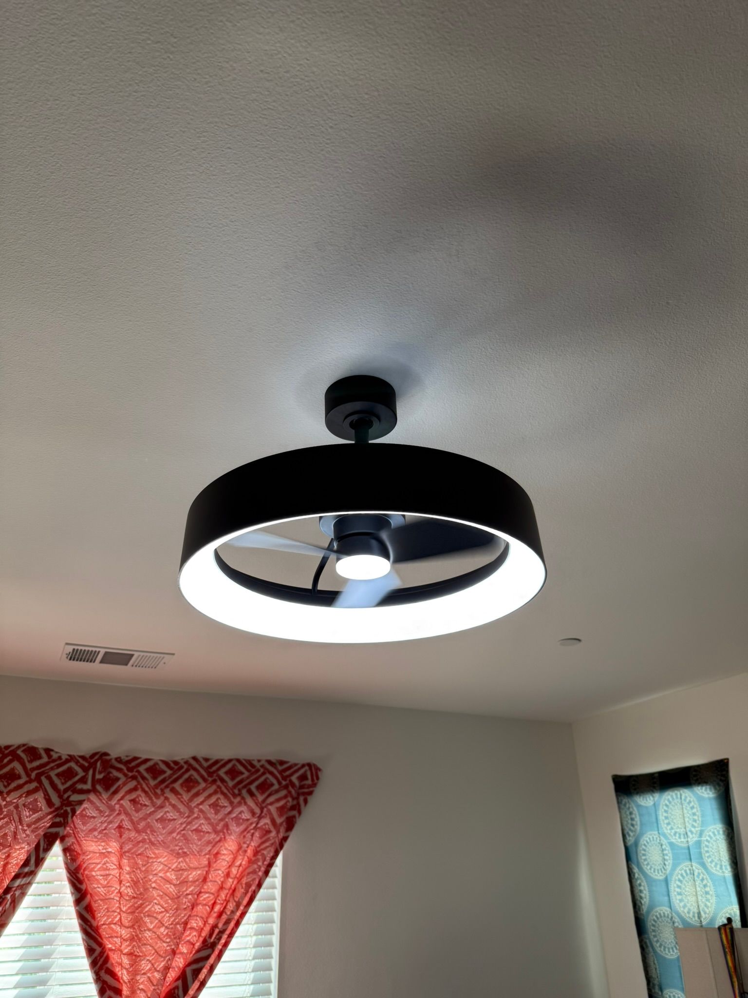Black ceiling fan with a white ring light, mounted on a white ceiling. Red patterned curtains on the left.