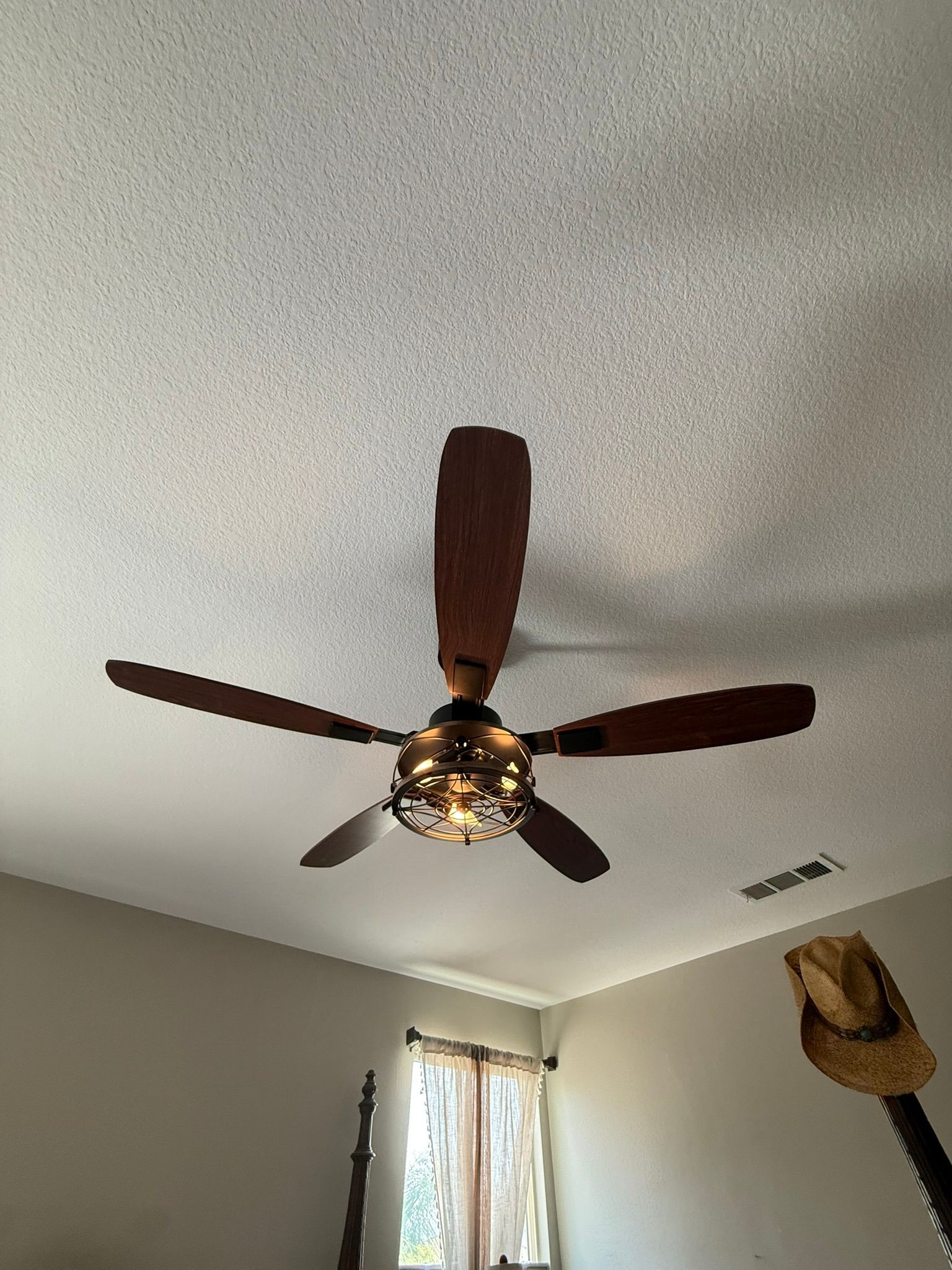 Ceiling fan with wooden blades and a decorative light fixture, in a room with a window and a straw hat.