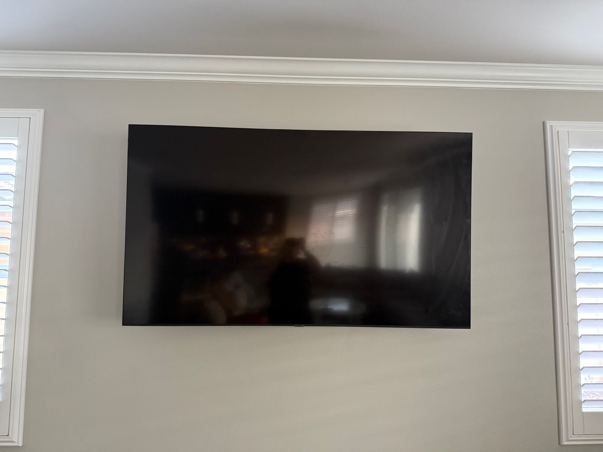 TV mounted on a light gray wall between two shuttered windows, beneath decorative molding.