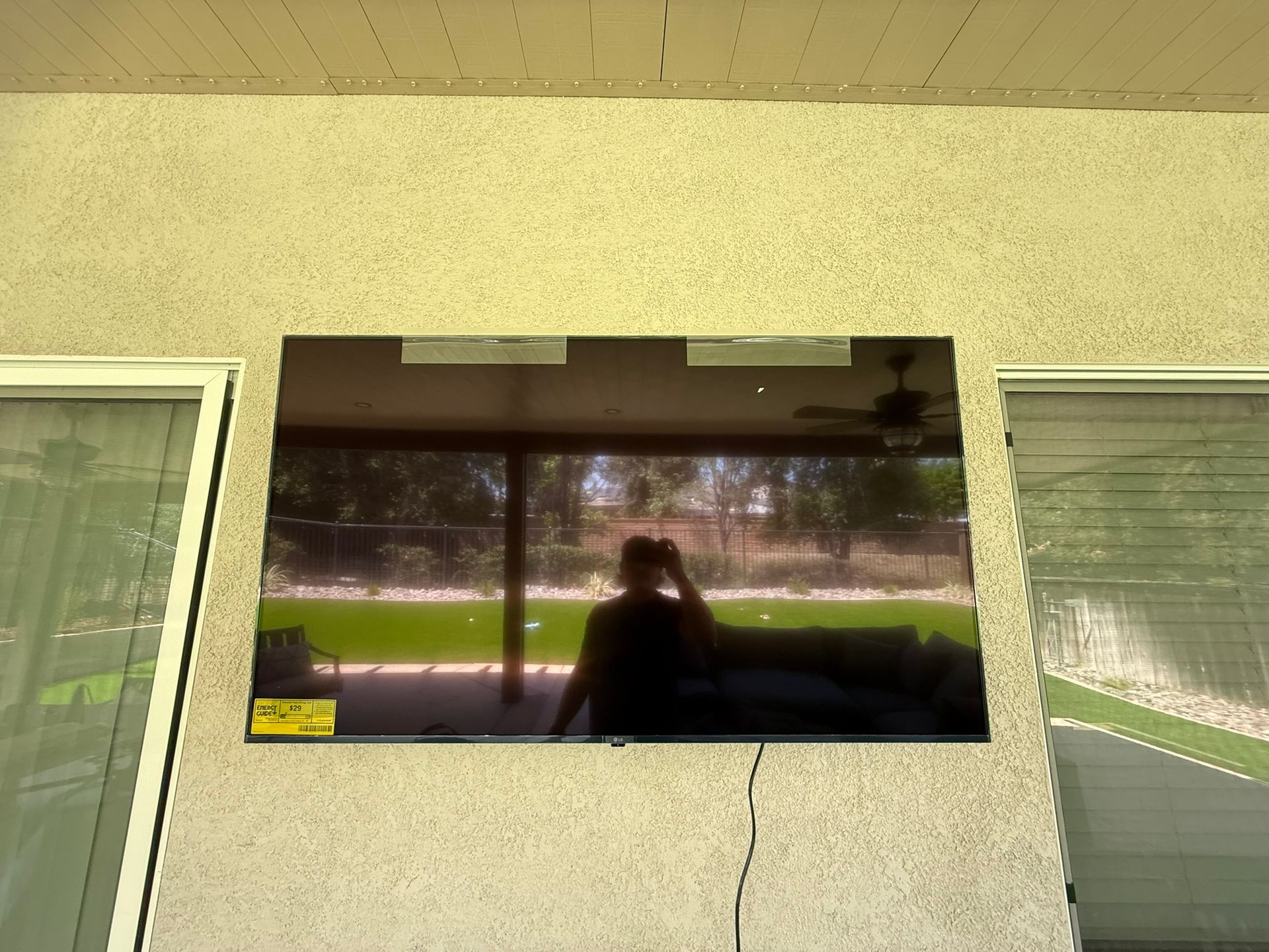 Television mounted on a stucco wall, reflecting a person taking a photo and outdoor scene.