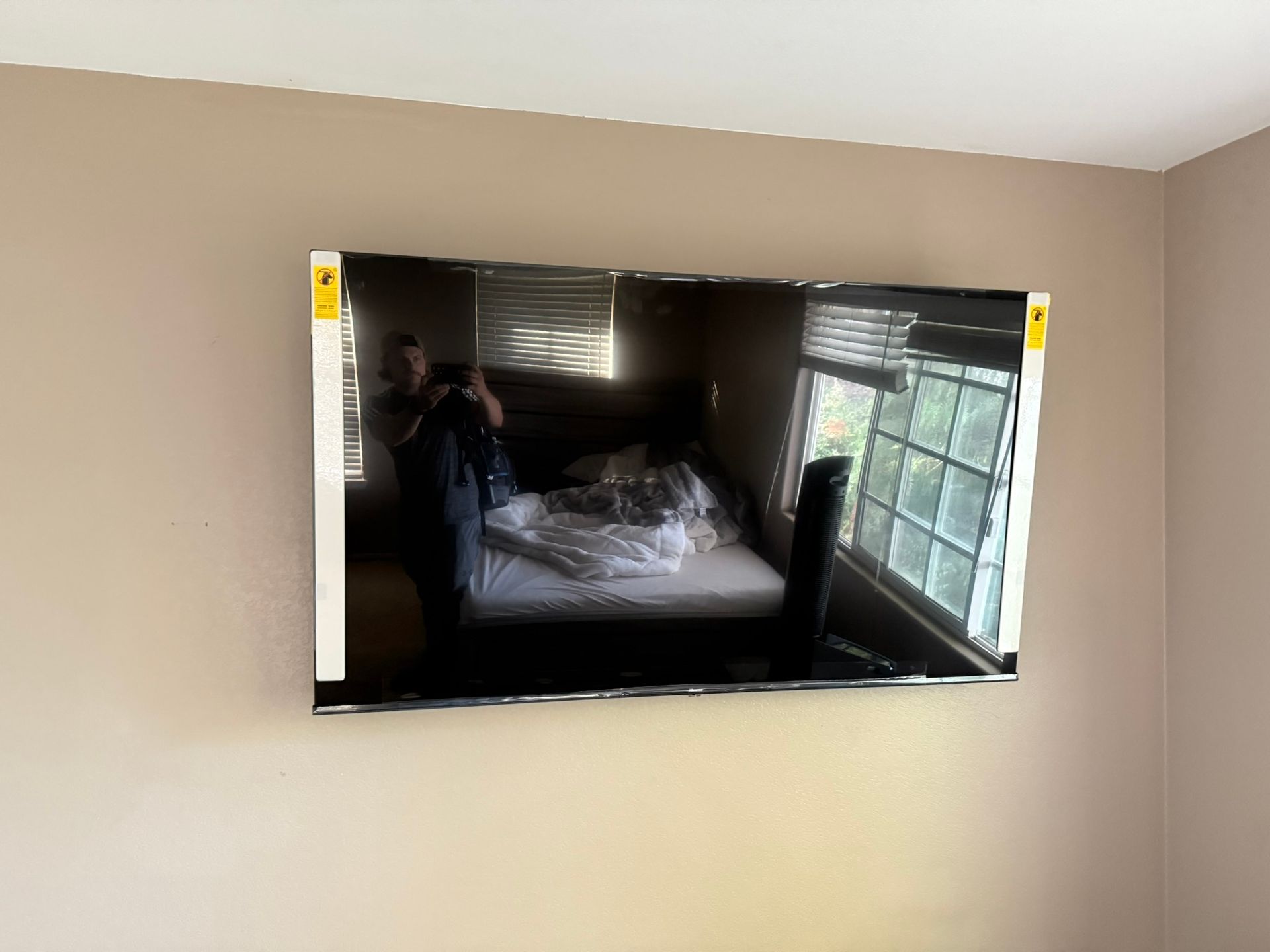 A large flat-screen TV mounted on a tan wall, reflecting a bedroom with a bed and windows.