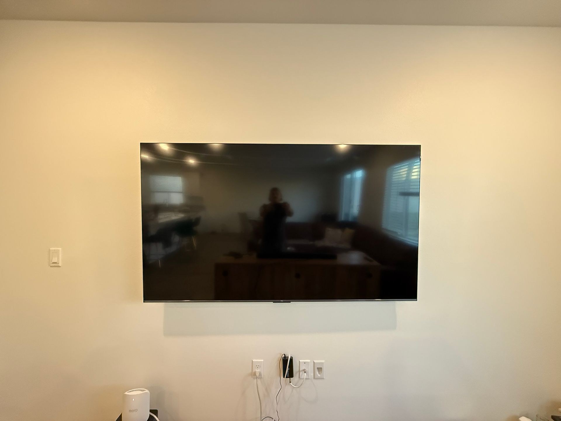 Large TV mounted on a beige wall, reflecting a room. White rectangular panel below the TV. Electrical outlets and cables visible.
