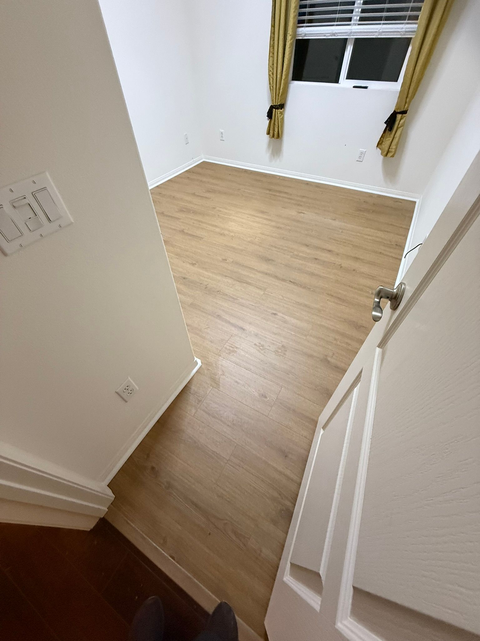 A room with wooden flooring, a window with curtains, and an open white door.