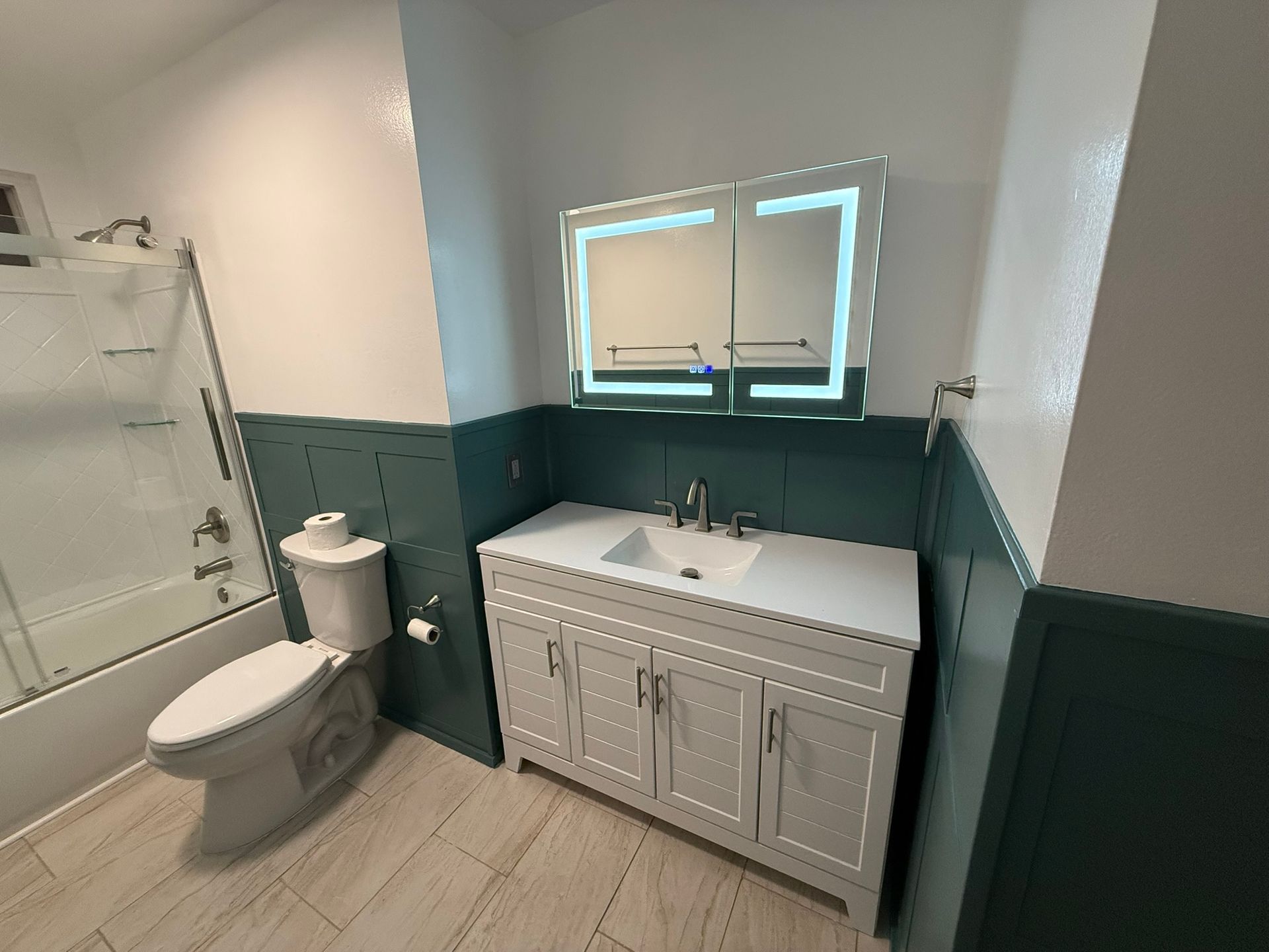 Bathroom with white vanity, toilet, and lit medicine cabinet against teal walls.