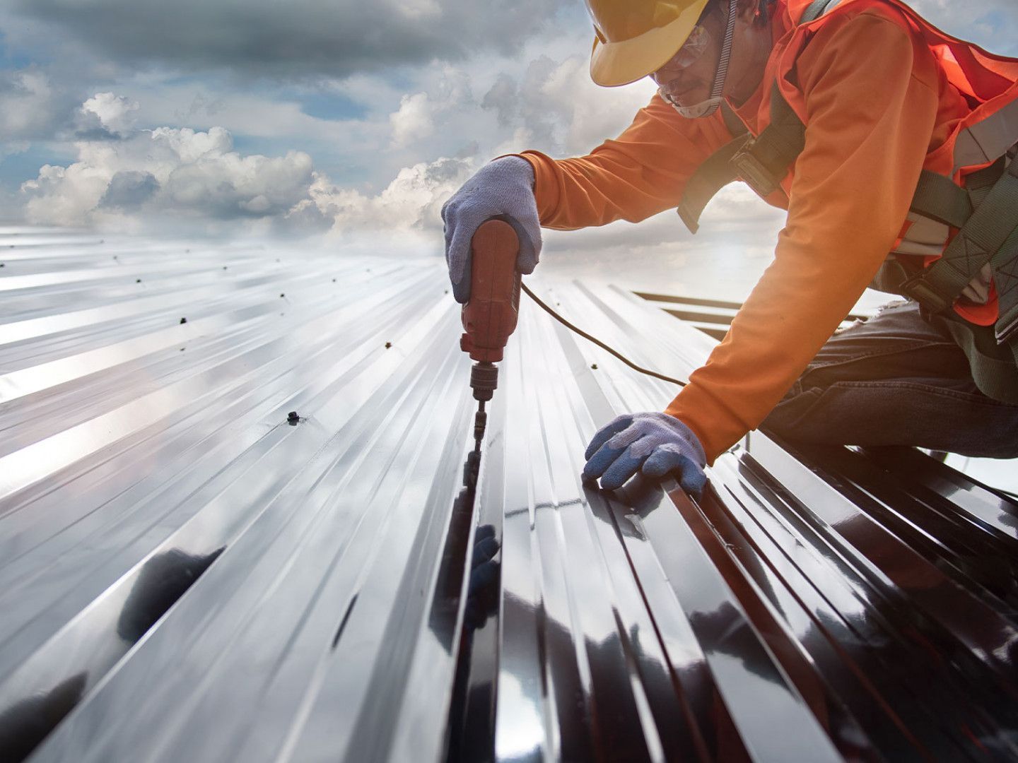 Roofer in safety gear drilling into a metal roof with a cloudy sky background.