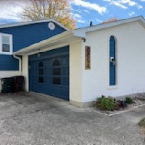 Blue and white house with a garage, driveway, and shrubs under a cloudy blue sky.