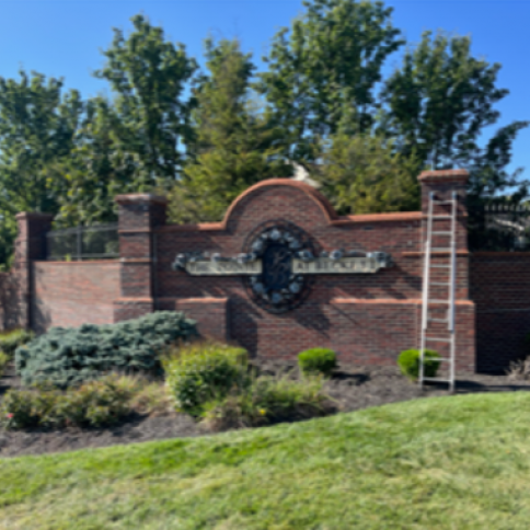 Brick entrance sign with a nameplate, trees in the background, ladder leaning on the side.