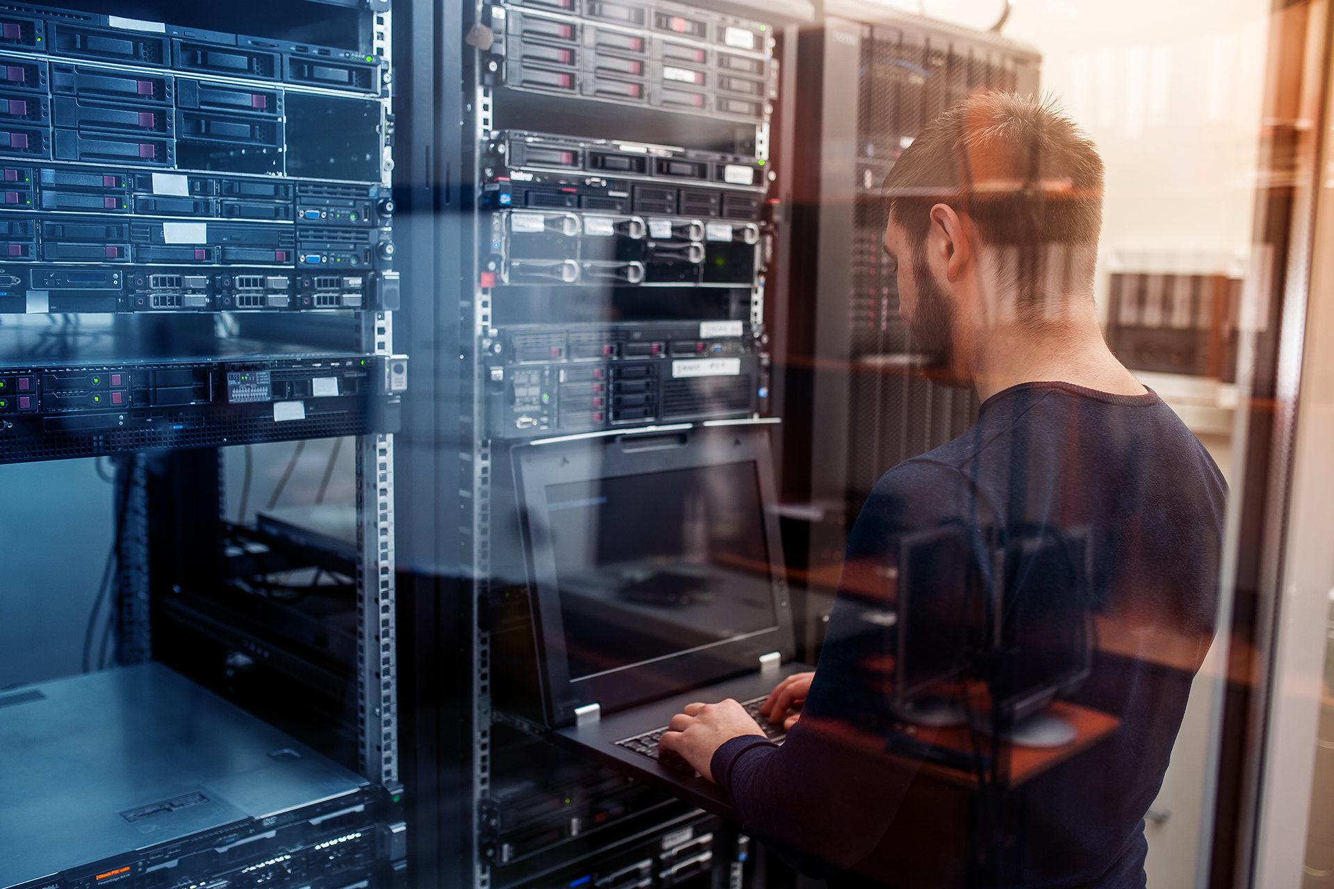 A man is working on a laptop in a server room.
