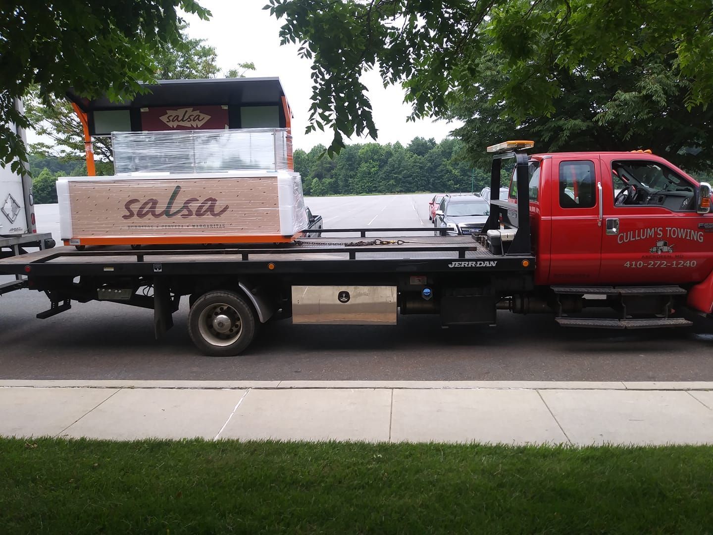 Red flatbed truck carrying wrapped furniture and boxes parked beside a sidewalk and trees
