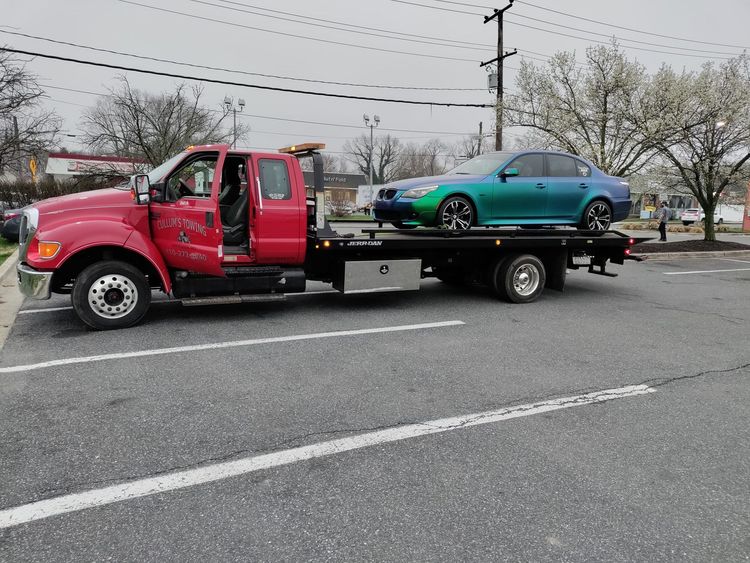 Red tow truck carrying a green hatchback on a wet roadside under gray skies Red tow truck carrying a green hatchback on a wet roadside under gray skies