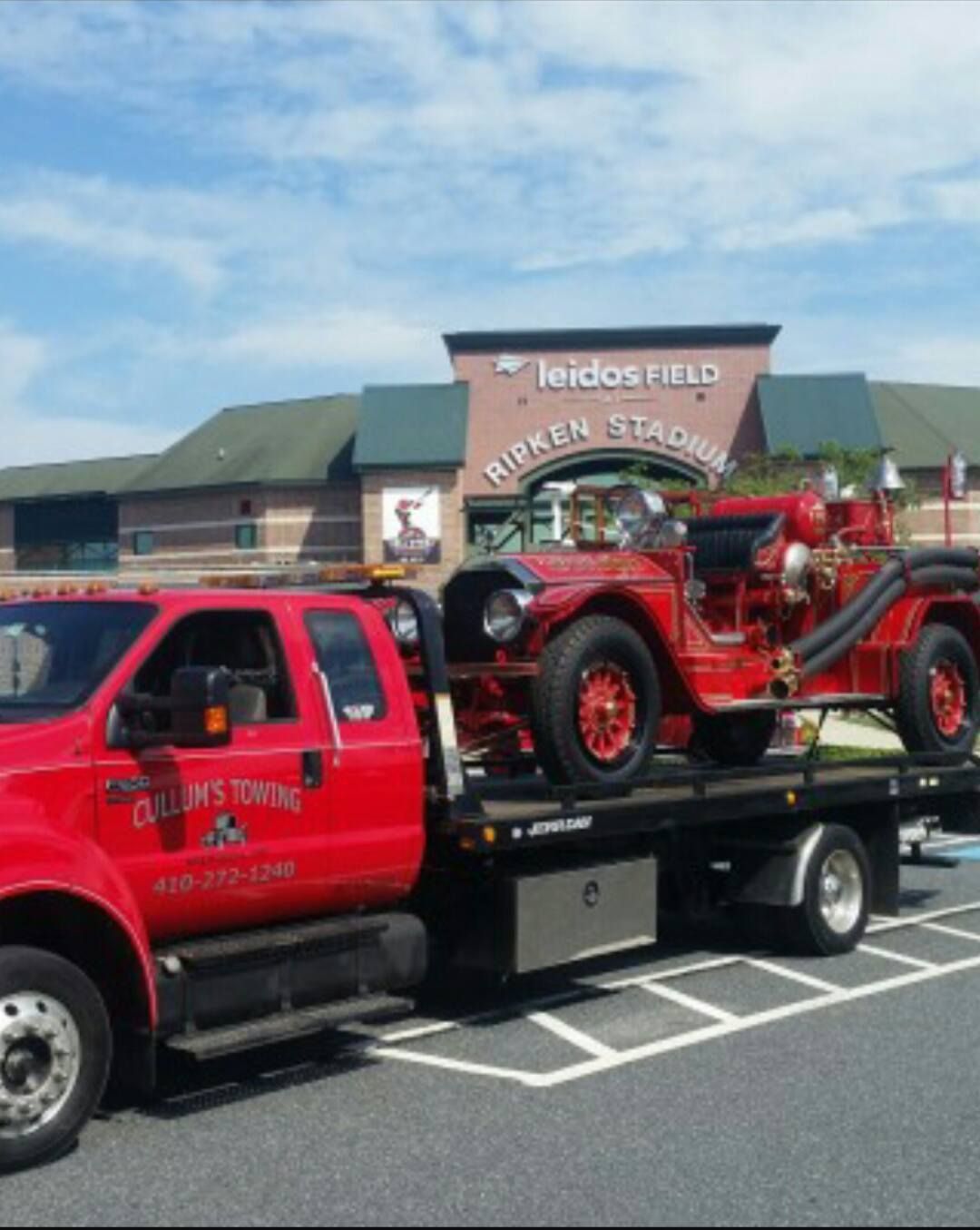 Red tow truck carrying a vintage red car in a parking lot outside a fairground building