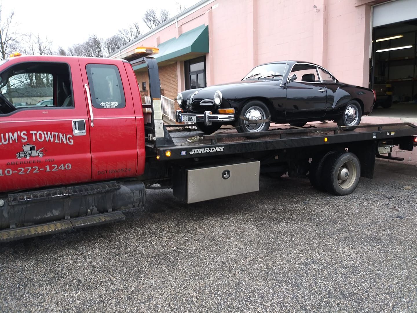 Red tow truck carrying a black sports car in a gravel lot near a garage