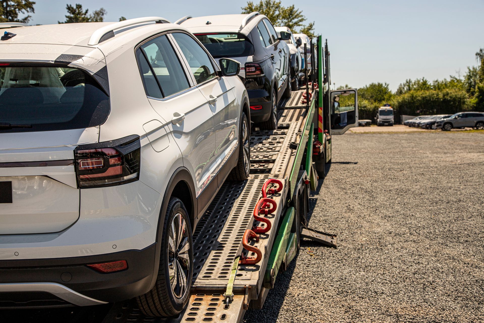 Cars loaded on a car carrier trailer in a gravel lot, with a white SUV in the foreground.