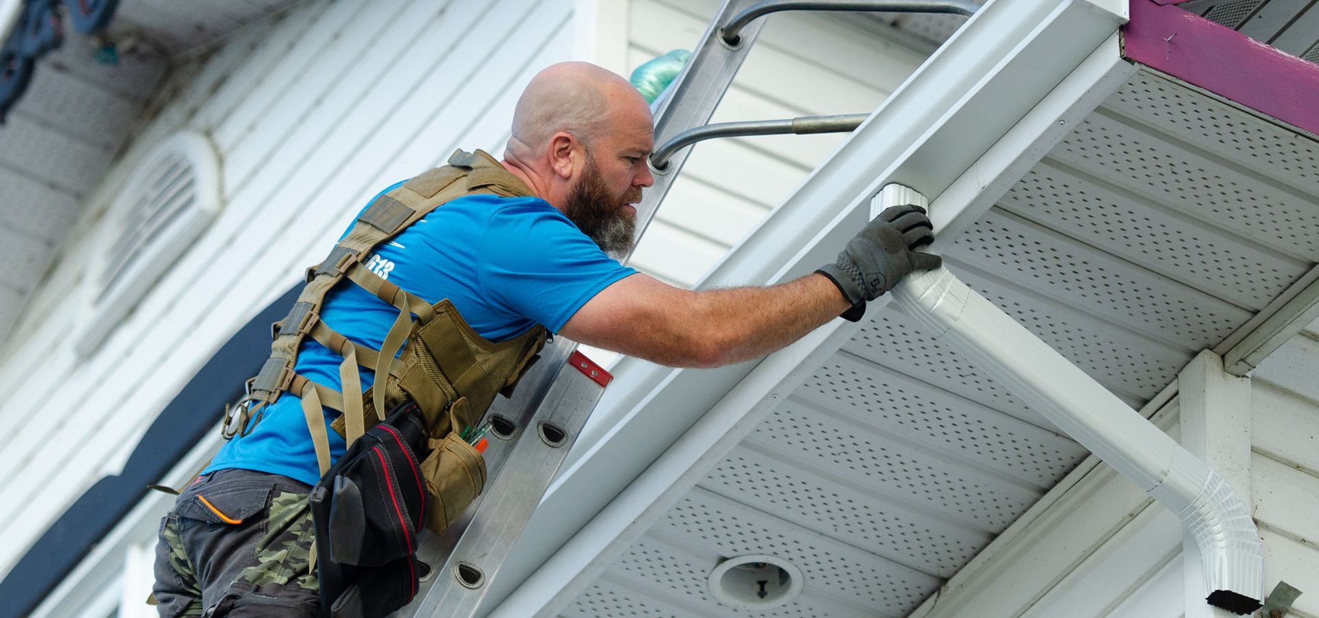 Un homme grimpe sur une échelle pour réparer une gouttière sur le côté d'une maison.