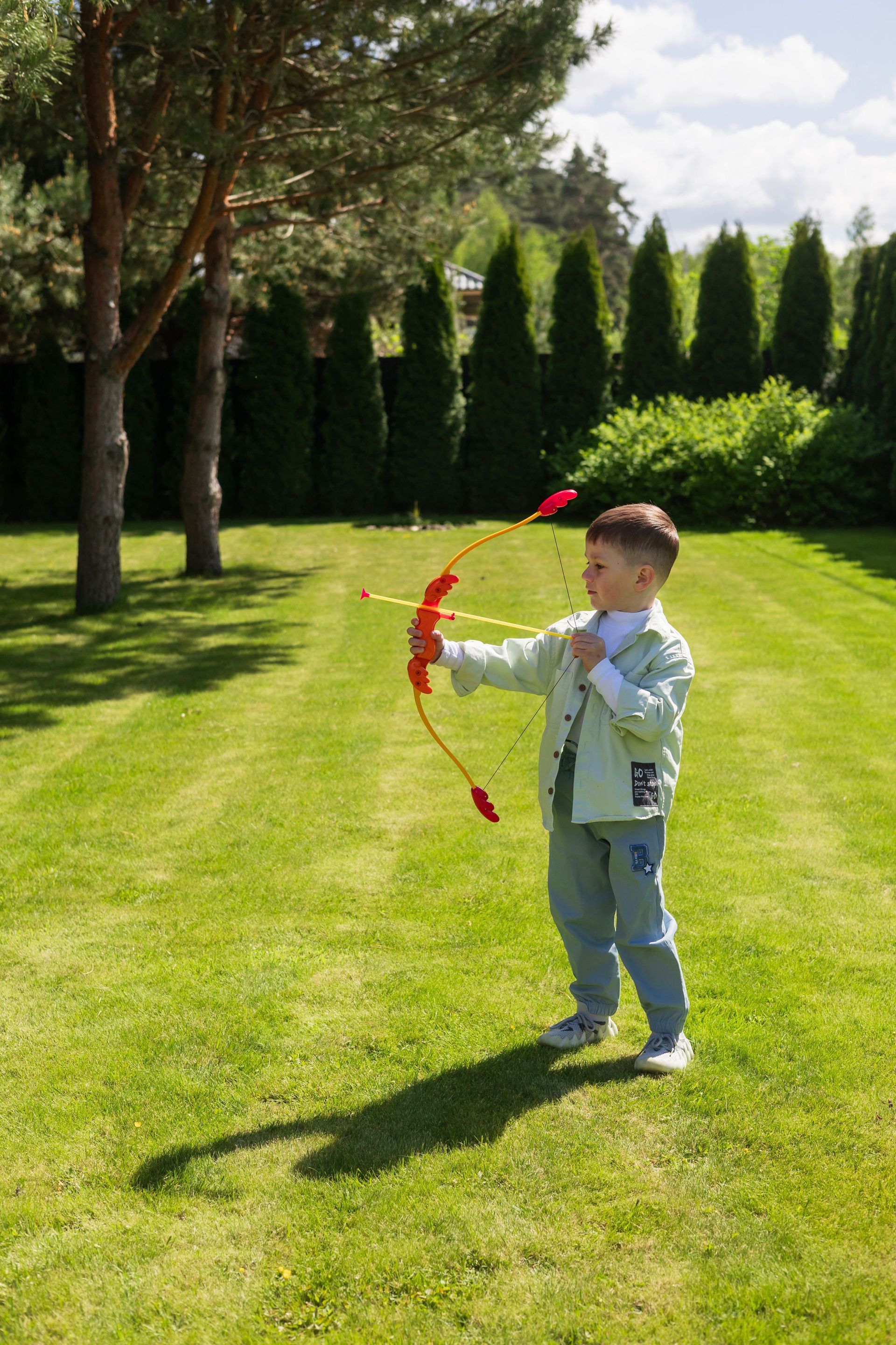 Child holding a red bow in a sunny green garden with trees and hedges
