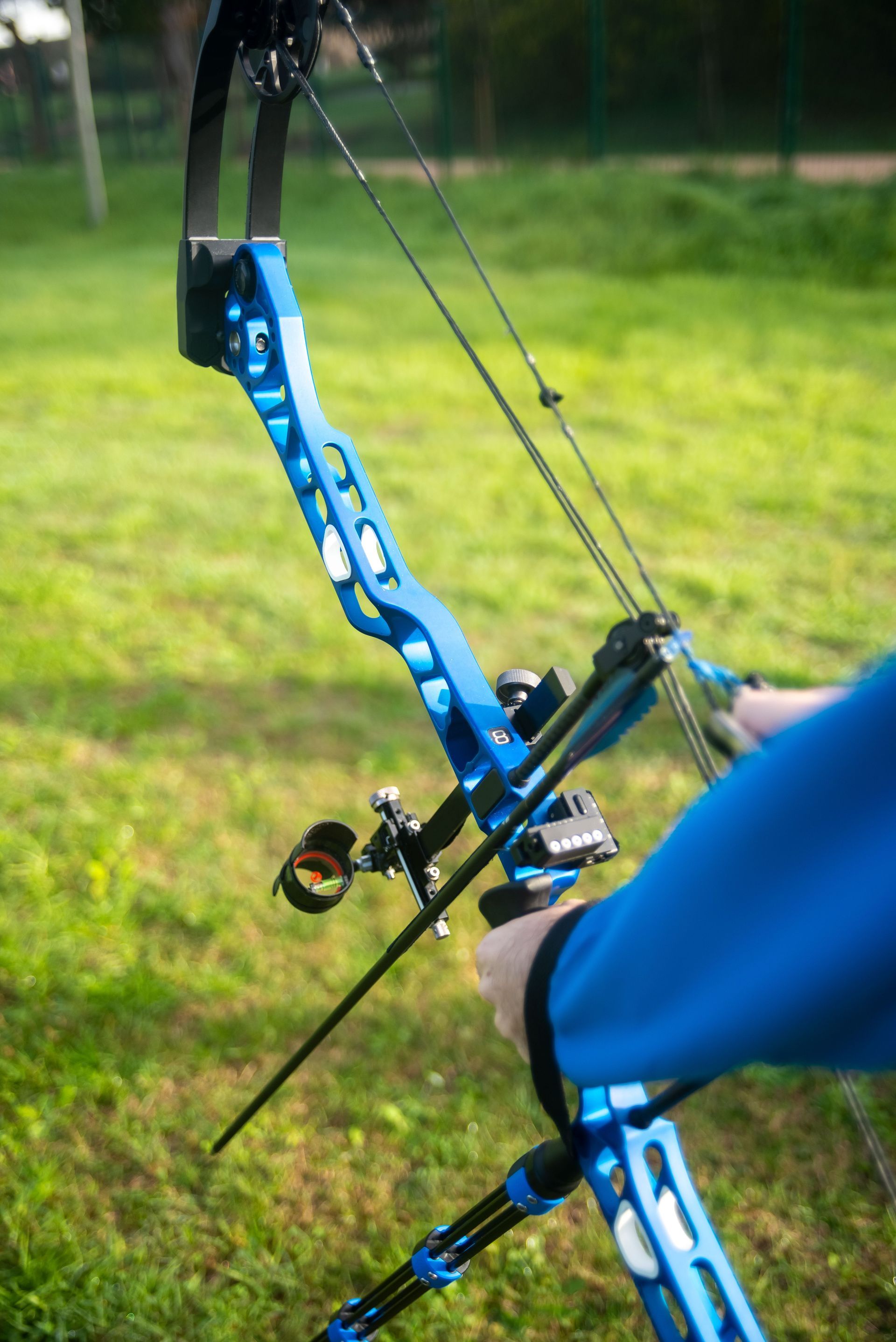 Person aiming a blue recurve bow in a grassy outdoor field