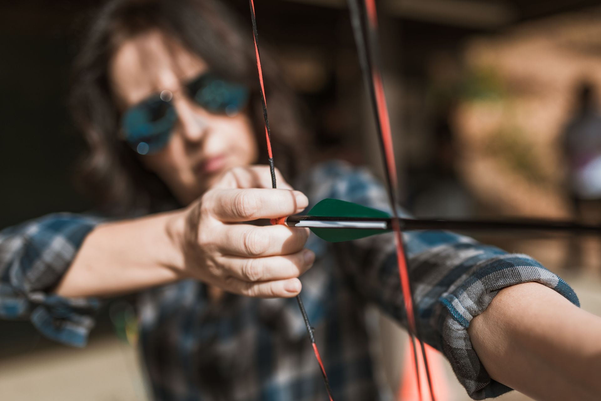 Person in plaid shirt aiming a bow and arrow, focused on the target outdoors.