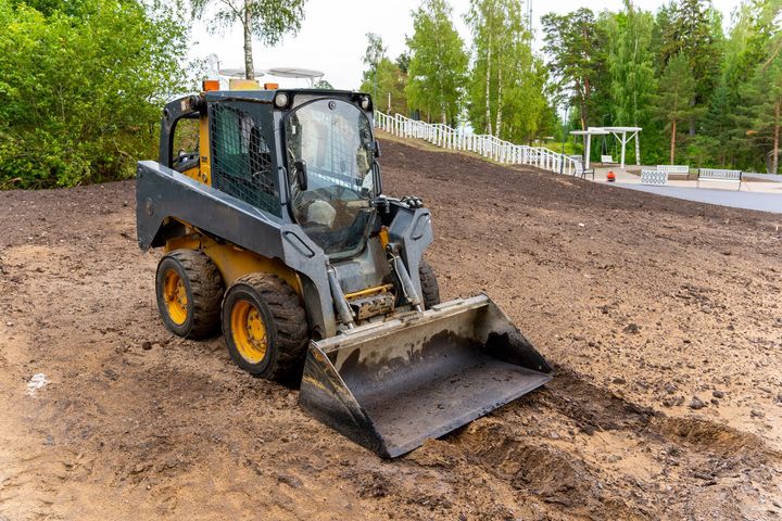 Compact loader leveling dirt at a construction site with trees in the background