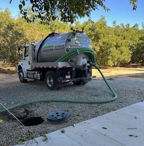 White septic service truck with green hose pumping at a roadside sewer opening under trees