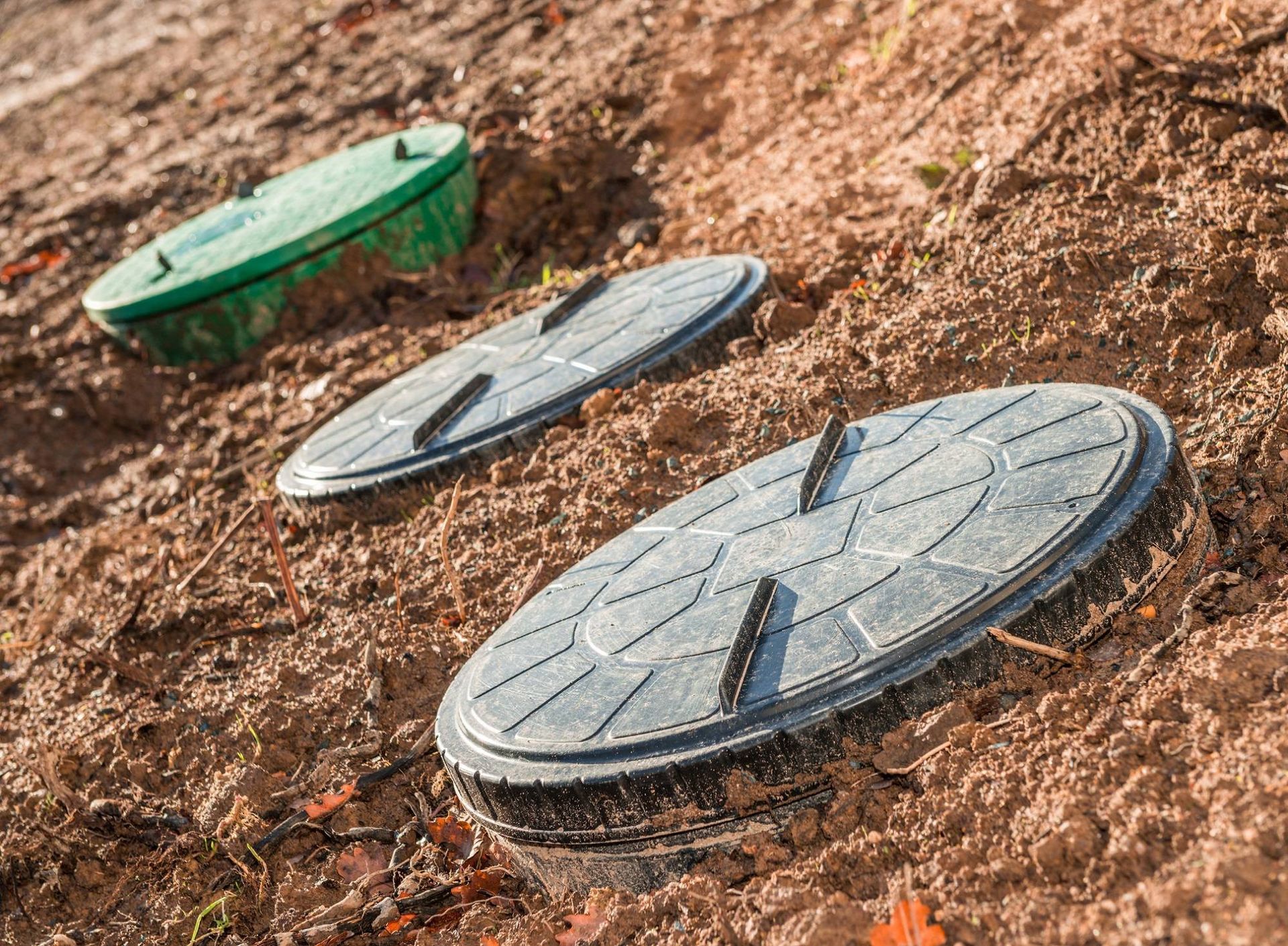 Three utility covers on a dirt slope, one green and two gray, partially buried in soil