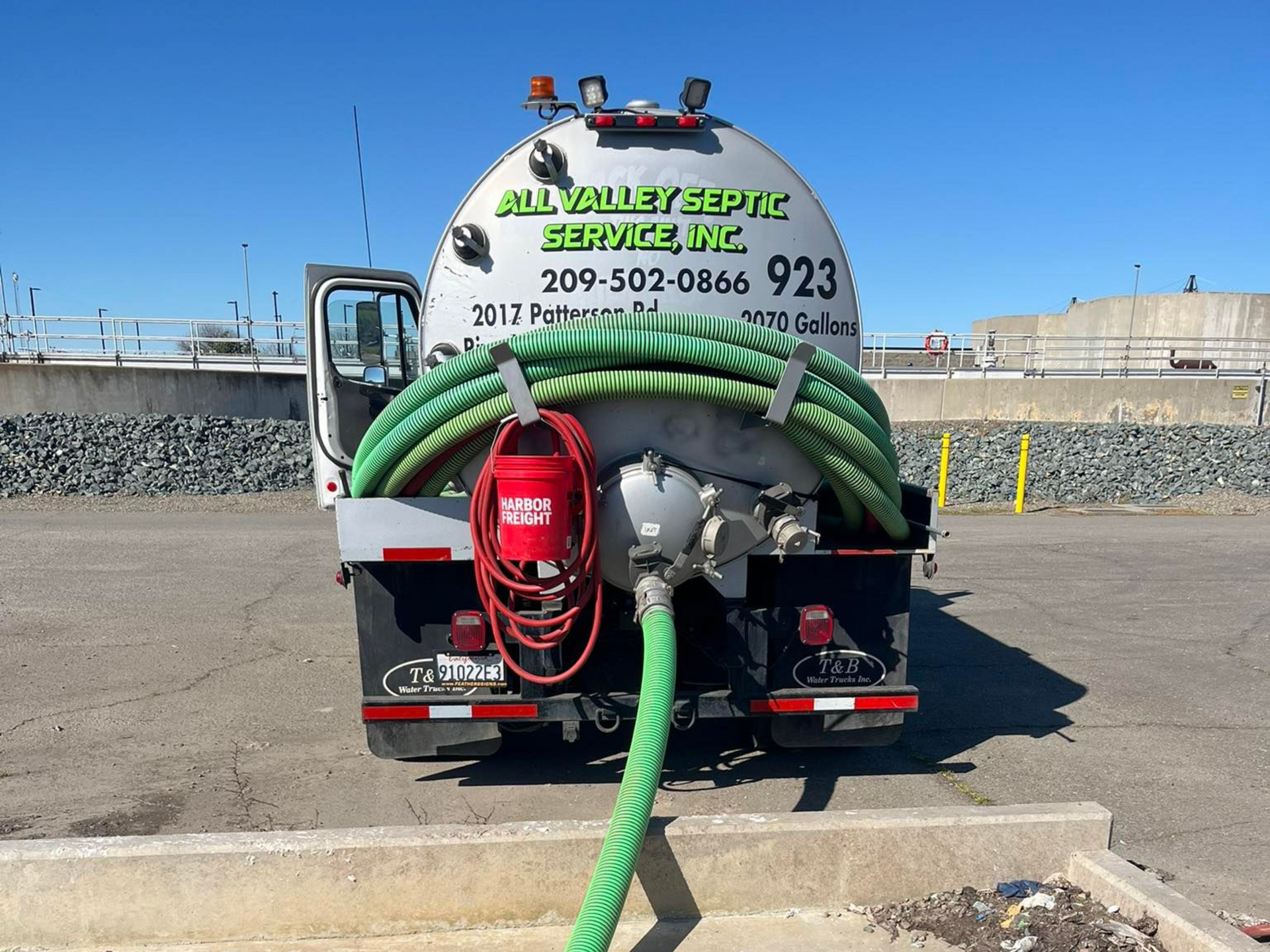 Vacuum truck with green hoses and red hose reel parked in an industrial lot under a blue sky