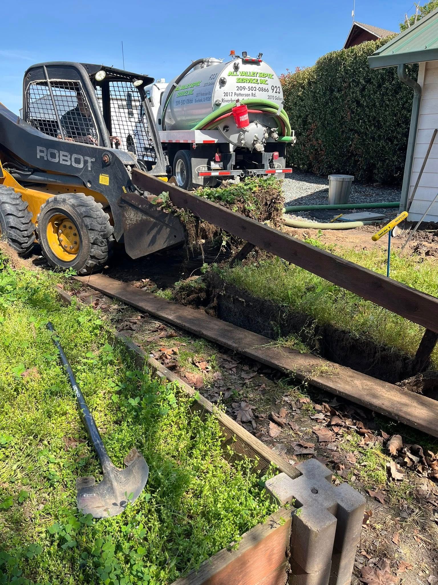 Black skid-steer loader moving wooden beams in a sunny yard with hedge and house in the background