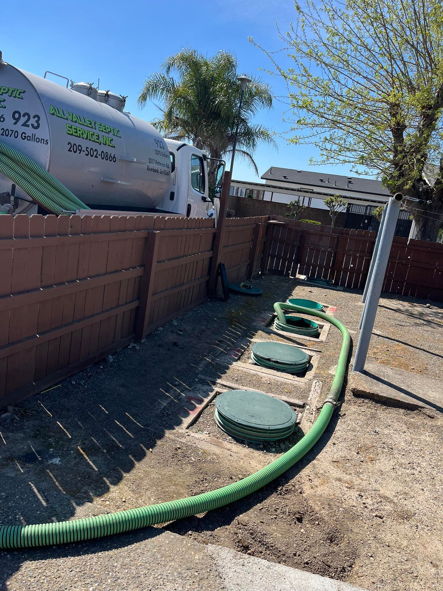 Backyard trench with green hose and round utility lids beside a fence and brick wall