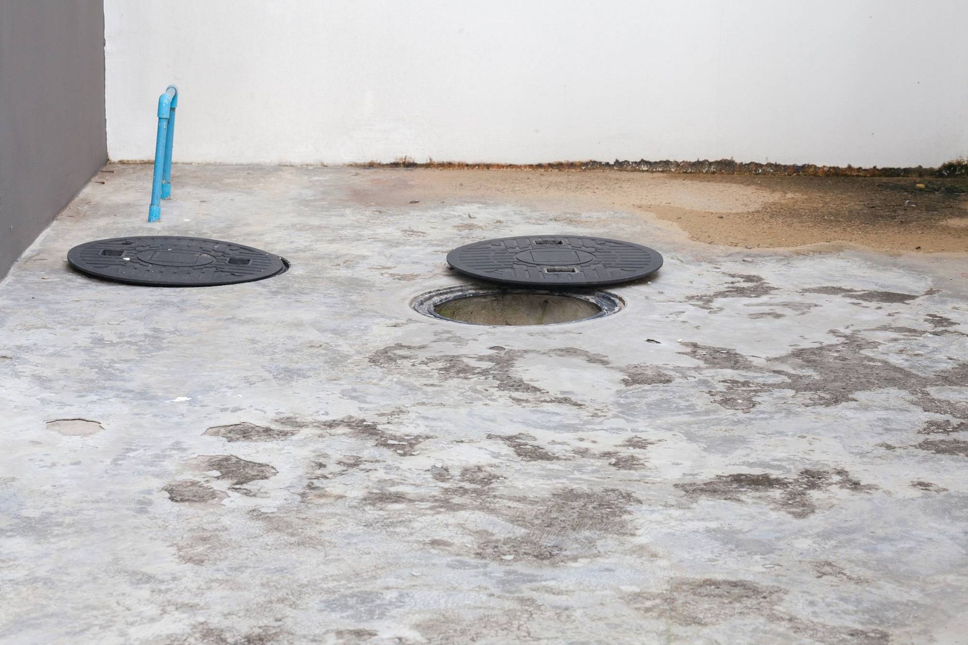 Two black manhole covers on a concrete surface, with a blue pipe near the wall.