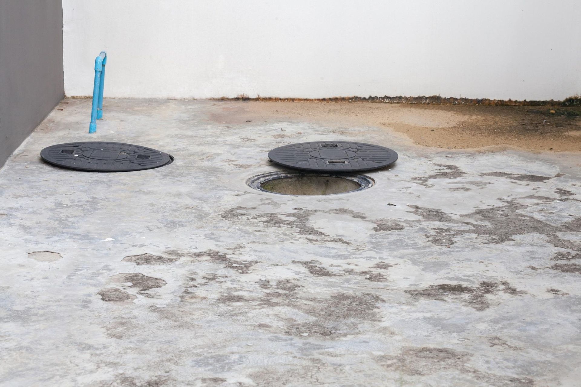 Outdoor concrete area with two round manhole covers and a blue pipe against a wall