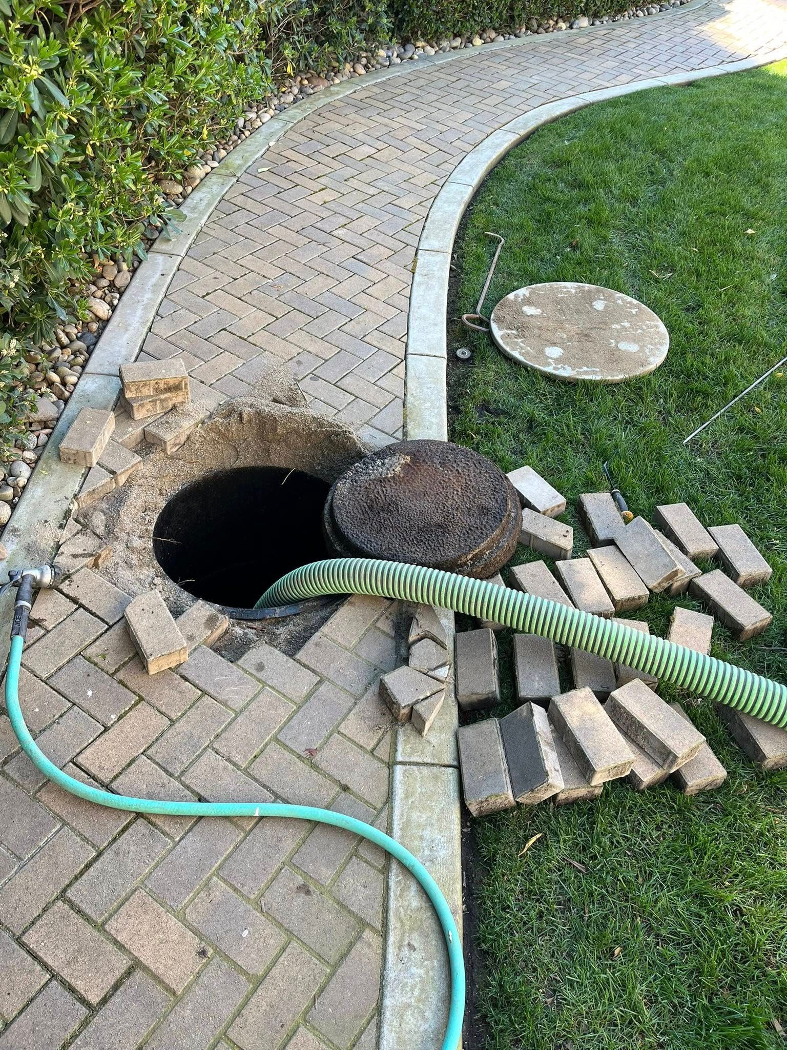 Clogged drainage hole beside a curved walkway, with loose bricks, a hose, and disturbed soil around it