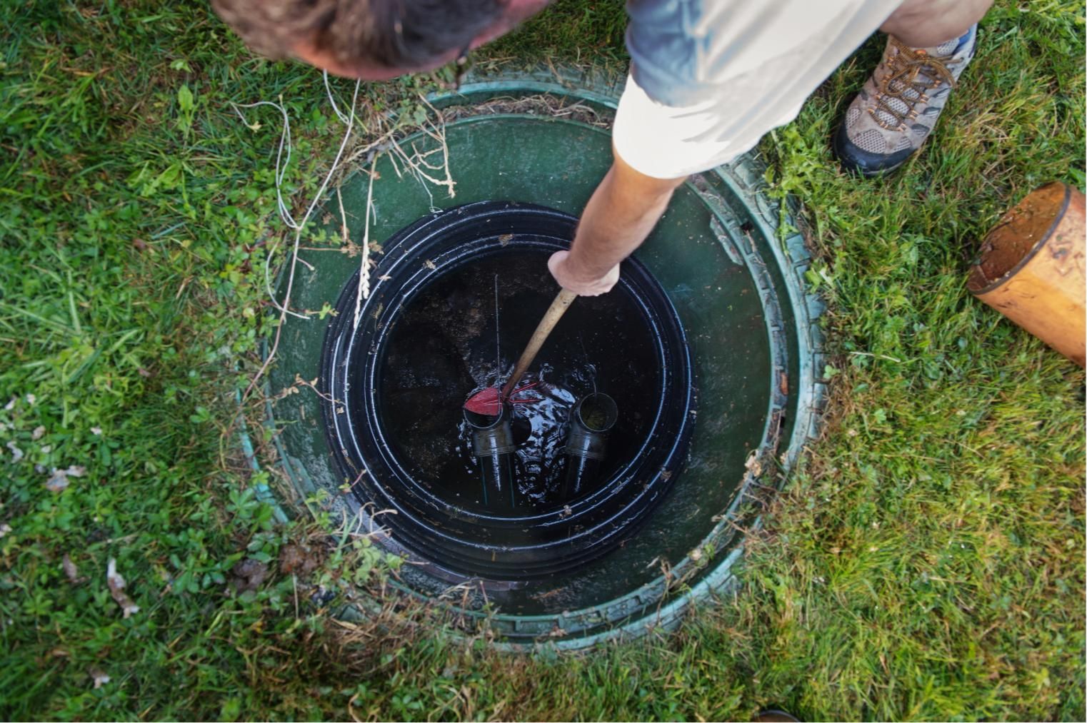 Person using a tool to clear a clogged drain in a grassy outdoor area.